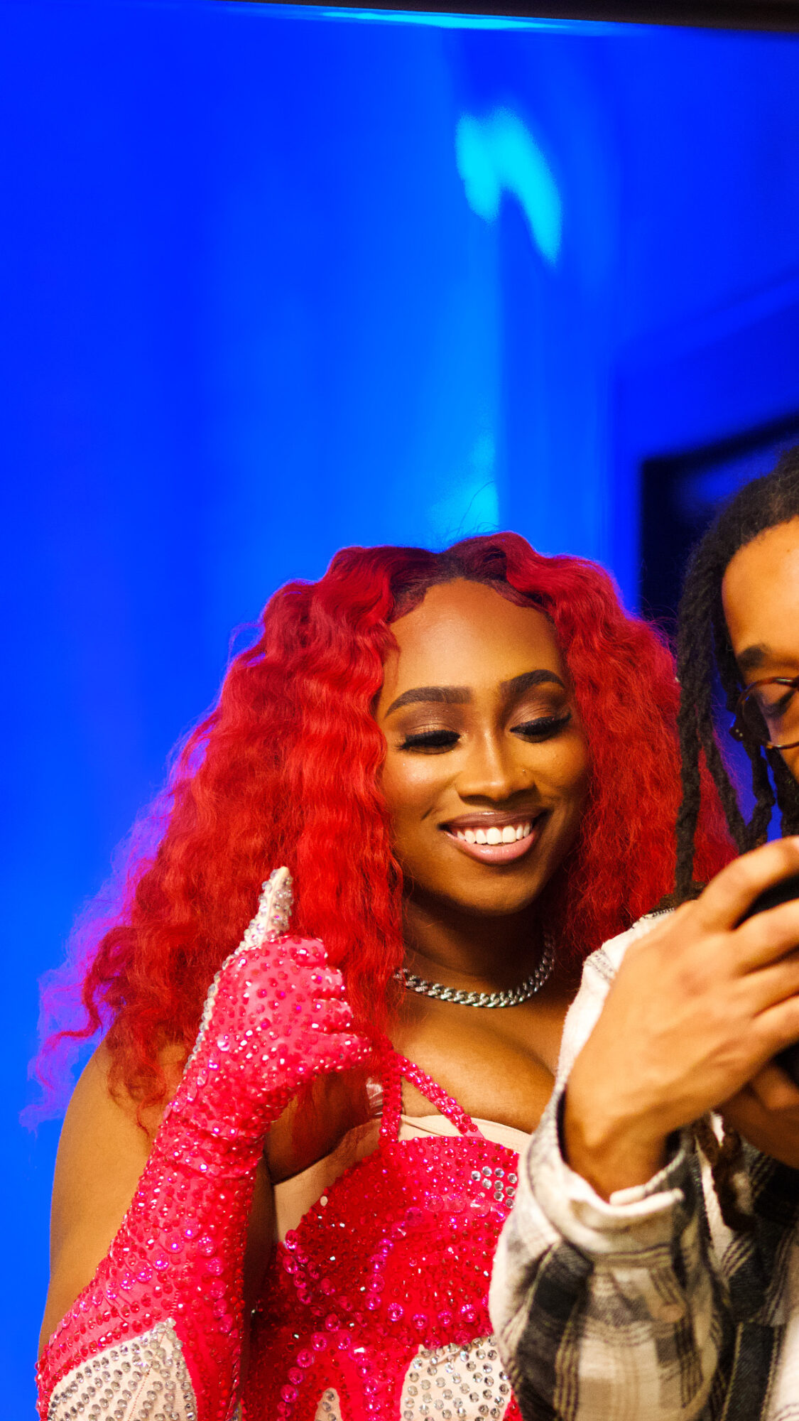 Smiling woman with red curly hair, wearing pink gloves and a necklace, in front of a blue background.