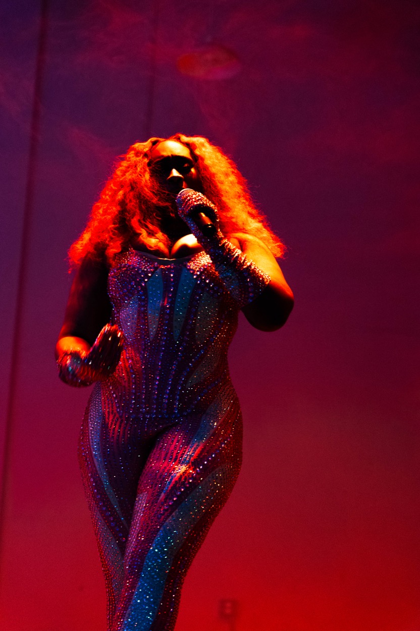 Performer with curly hair singing into a microphone on stage, illuminated by red and purple lighting.