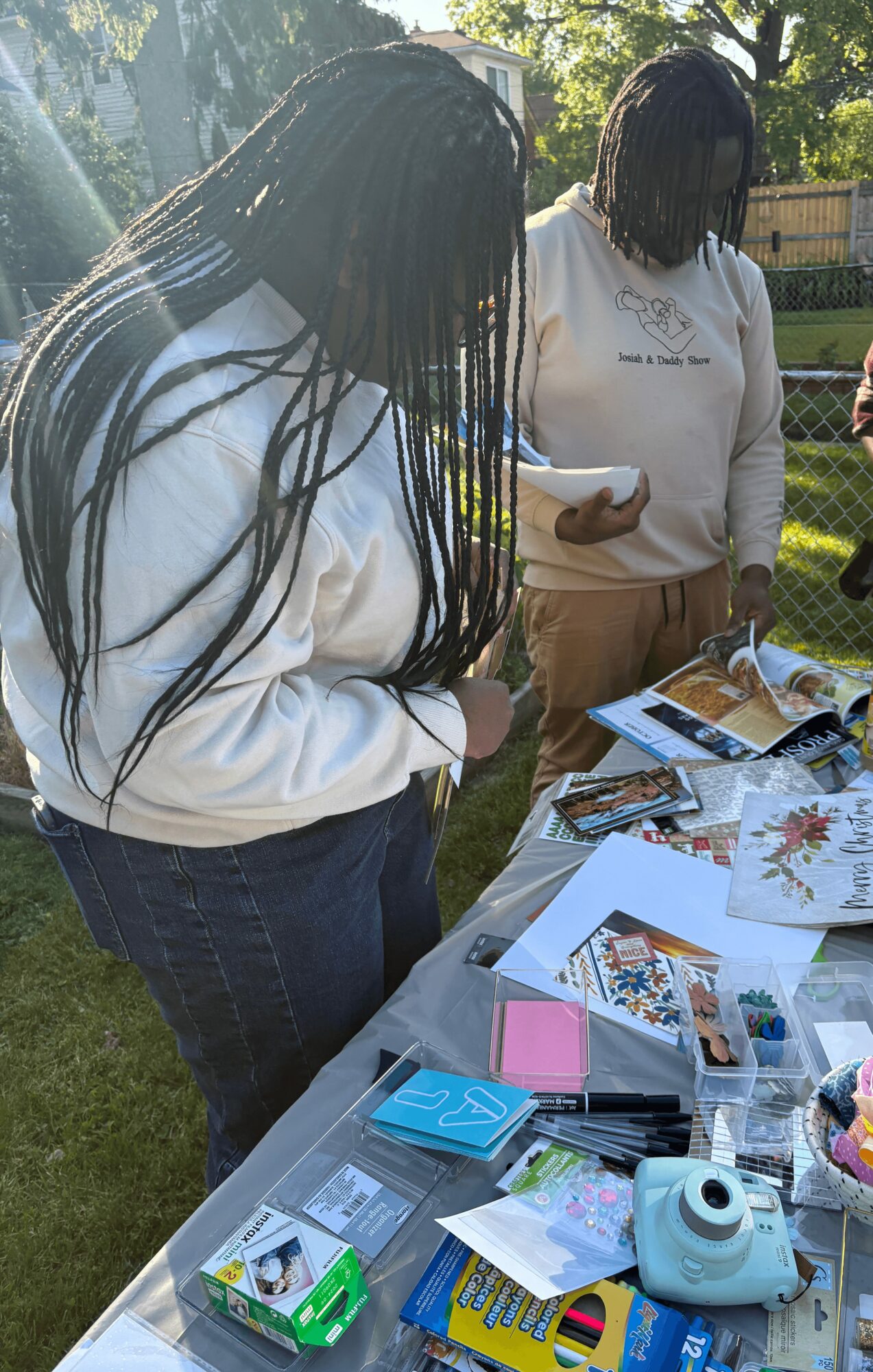 Two people with long hair looking at a table with various papers and items outdoors, trees and a fence in background.