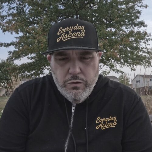 Man wearing a black cap and jacket outdoors, with trees and houses in the background.