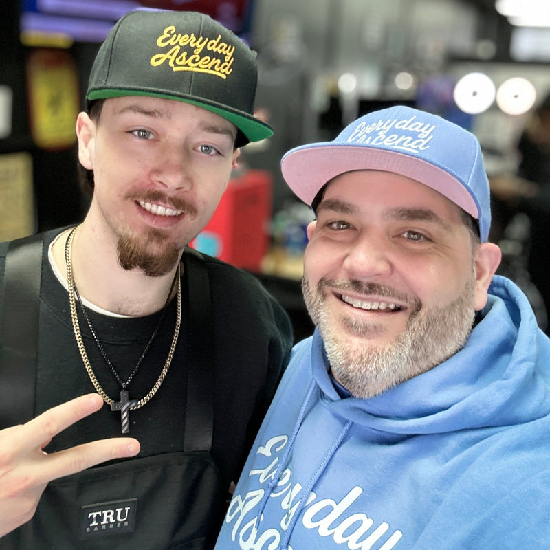 Two men smiling, wearing caps with text, one in black and the other in blue hoodie, indoors with blurred background.