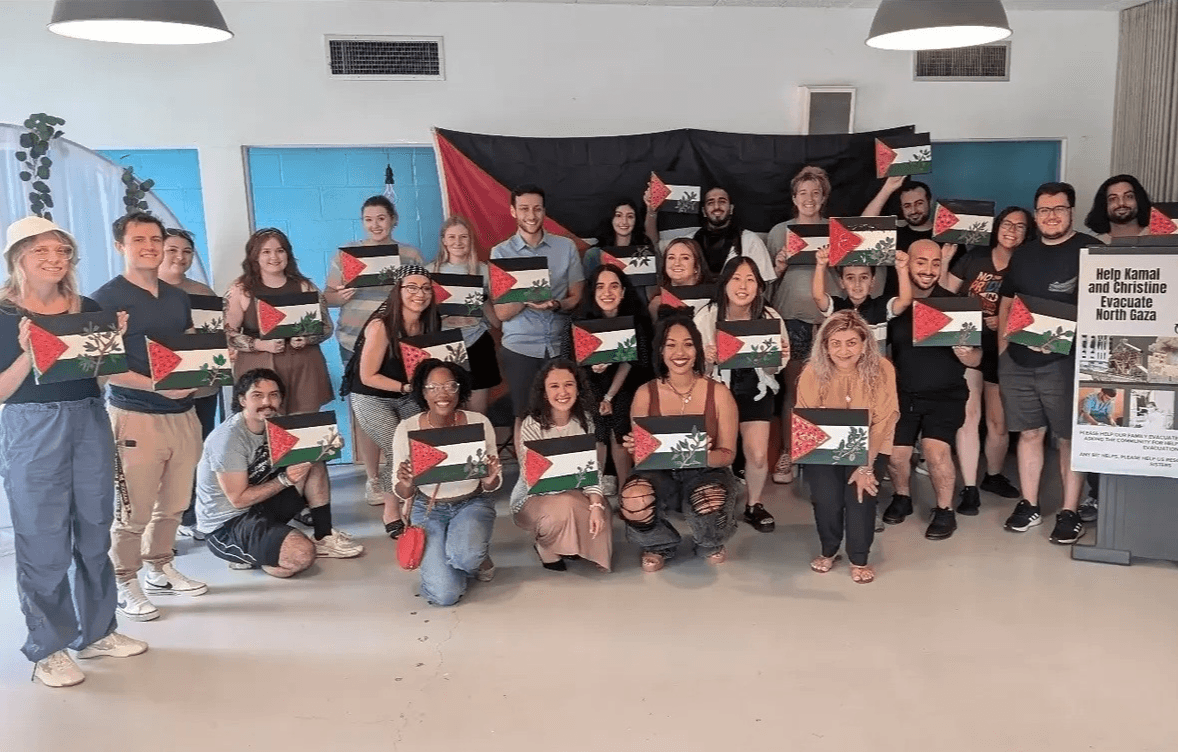 Group of people holding flags, posing indoors with a black and red backdrop, some kneeling and some standing.