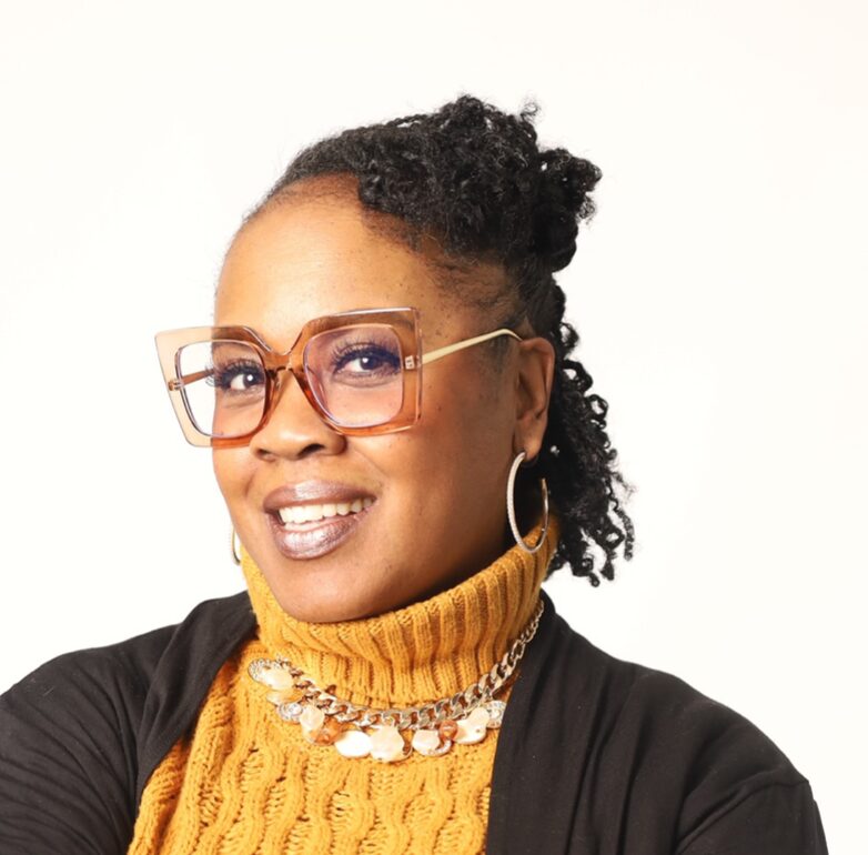 Woman with glasses, curly hair, wearing a yellow turtleneck and layered necklaces, smiling at the camera.