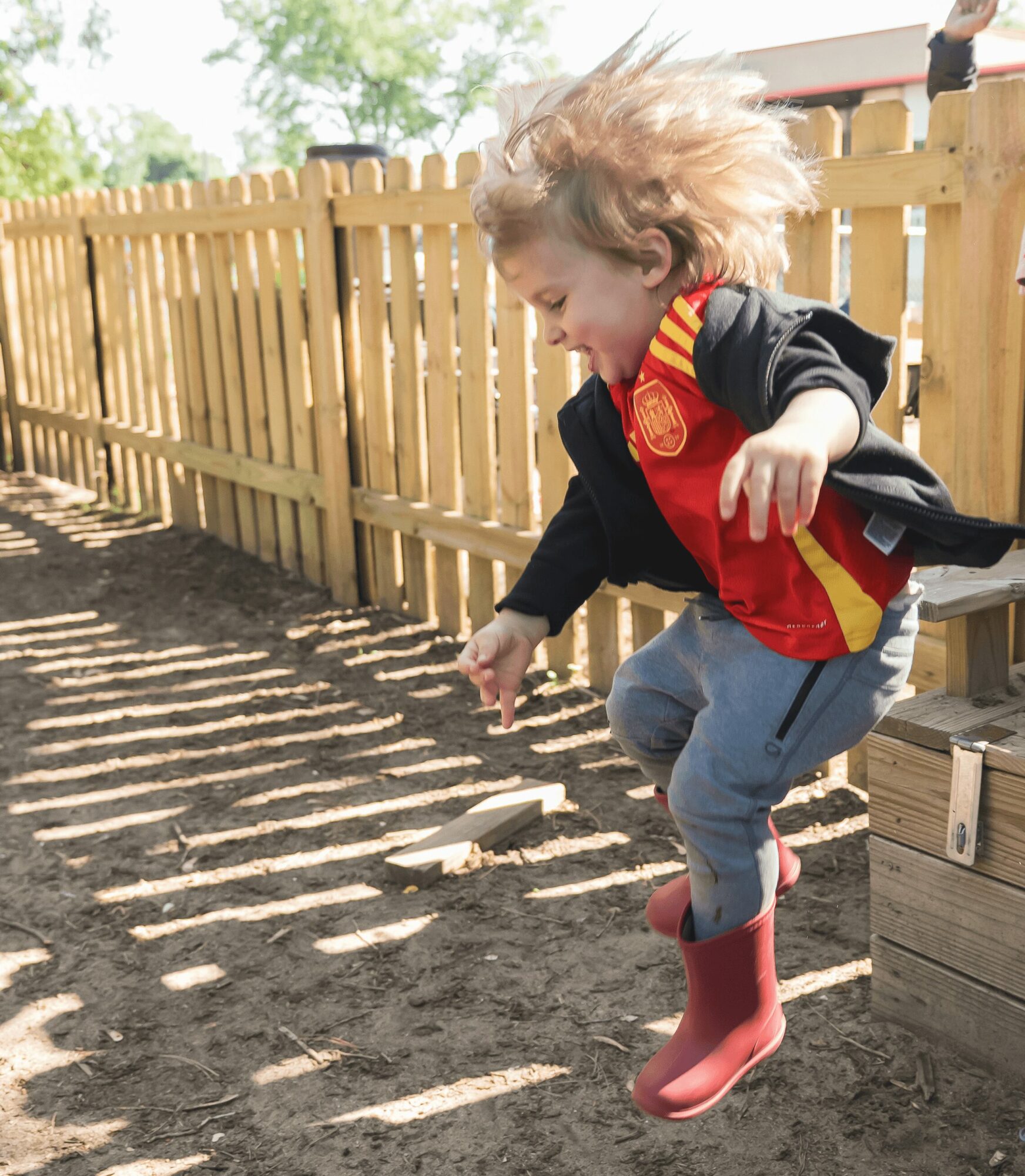 Child jumping outdoors near a wooden fence, with sunlight casting shadows on the ground, wearing red rain boots and a jacket.