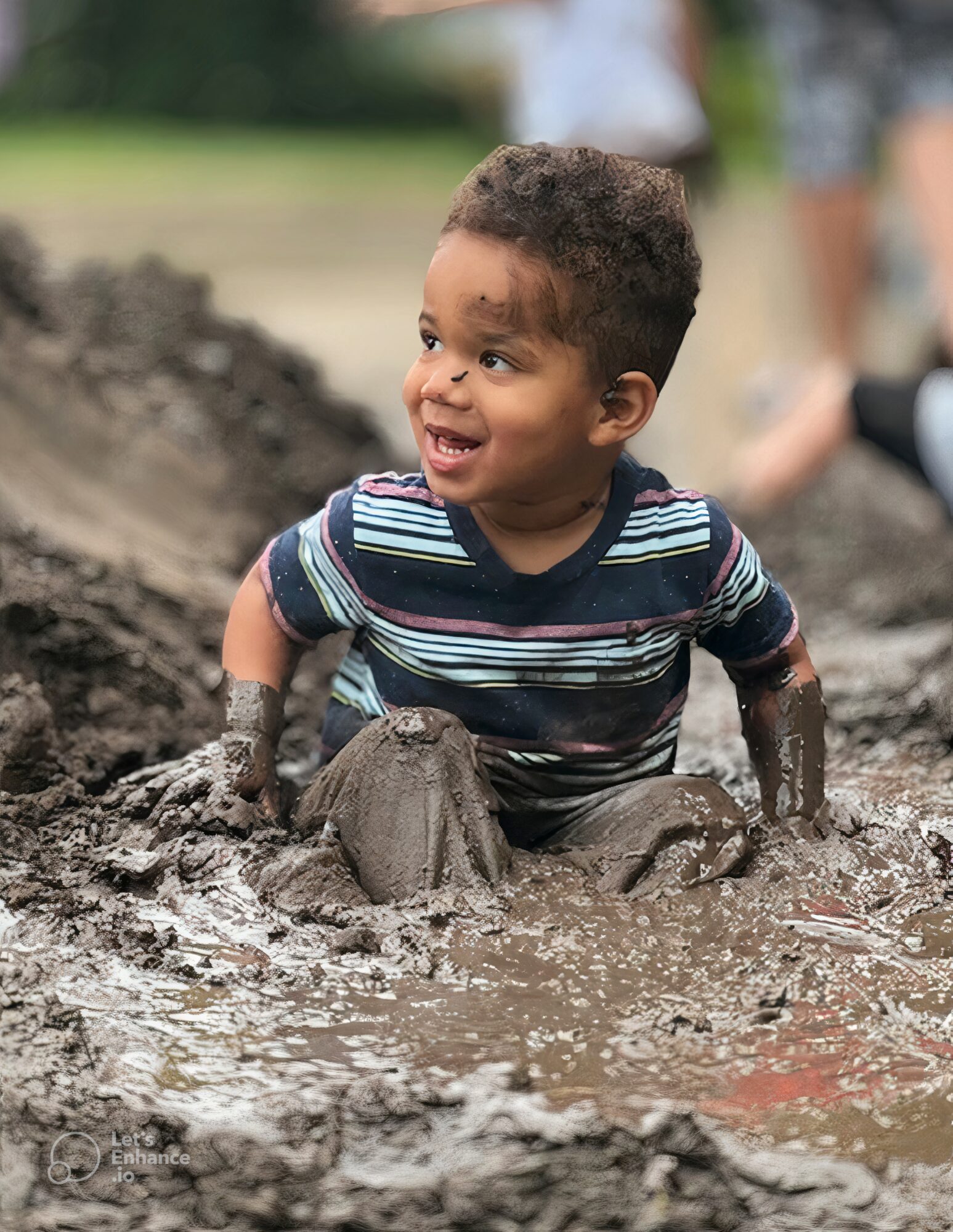 Young child with curly hair smiling while playing in muddy water outdoors, wearing a striped shirt.