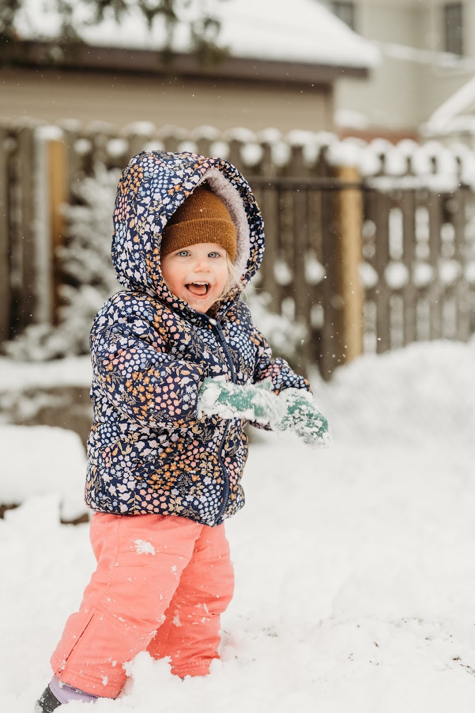 Child in winter coat and hat playing in snow, smiling, holding a snowball, outdoors with a wooden fence and snow-covered ground