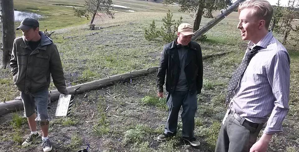 Three men standing outdoors near a fallen tree, with grass and trees in the background.