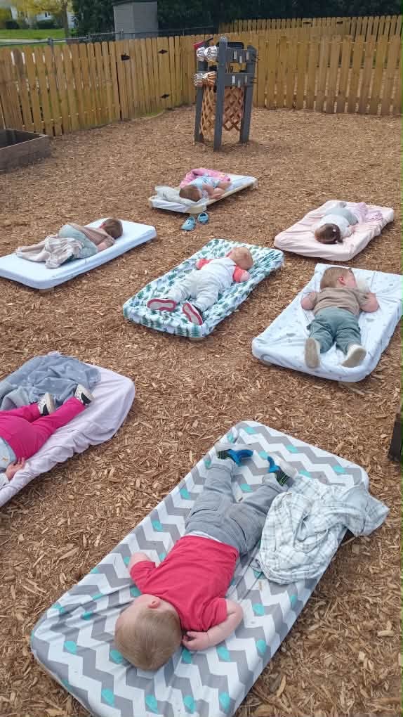Six babies lying on mats outdoors in a fenced area, some sleeping, some awake, with a wooden play structure in background.