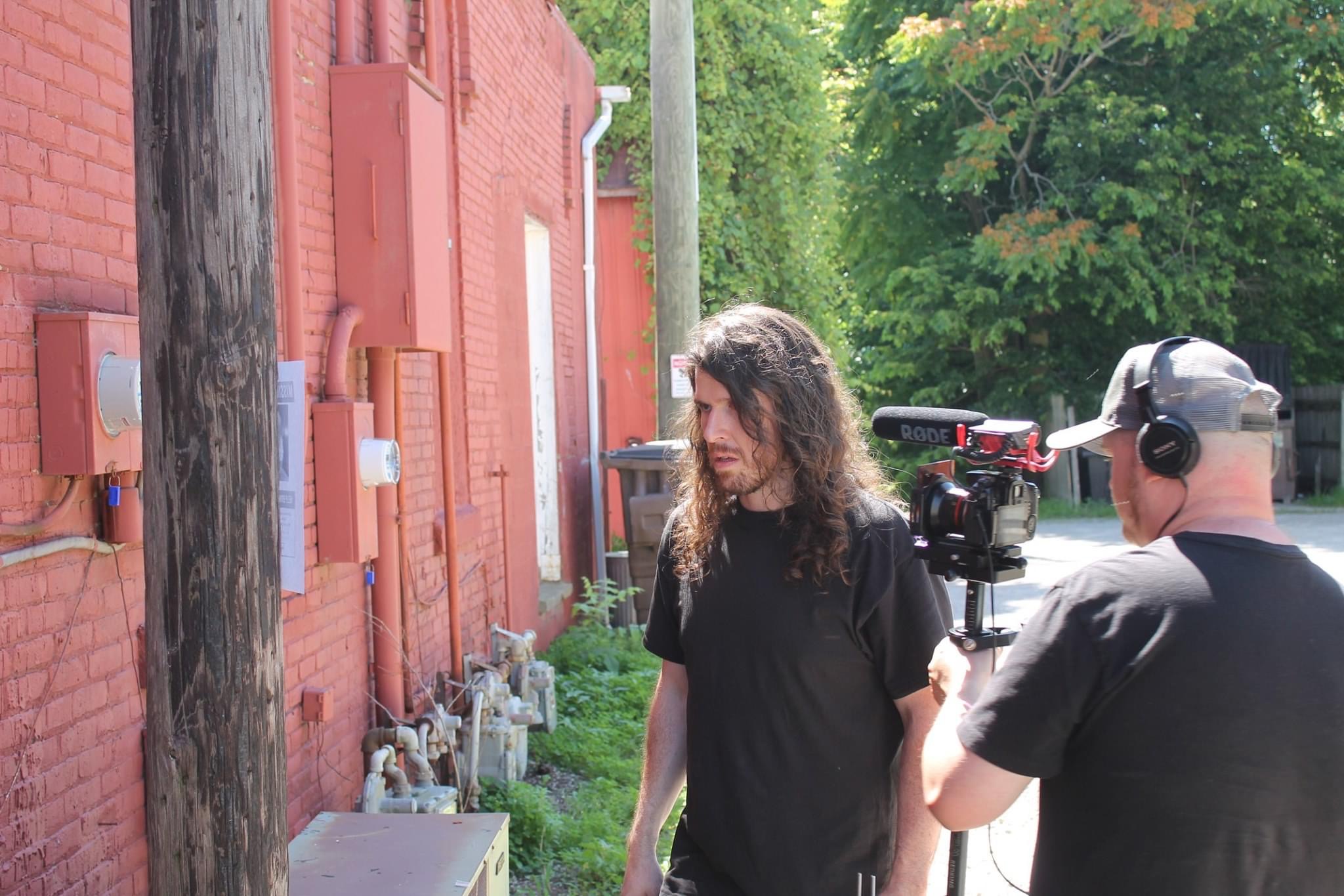 Two men outdoors near a red brick building, one with long hair and the other with a cap and headphones, filming with a camera.