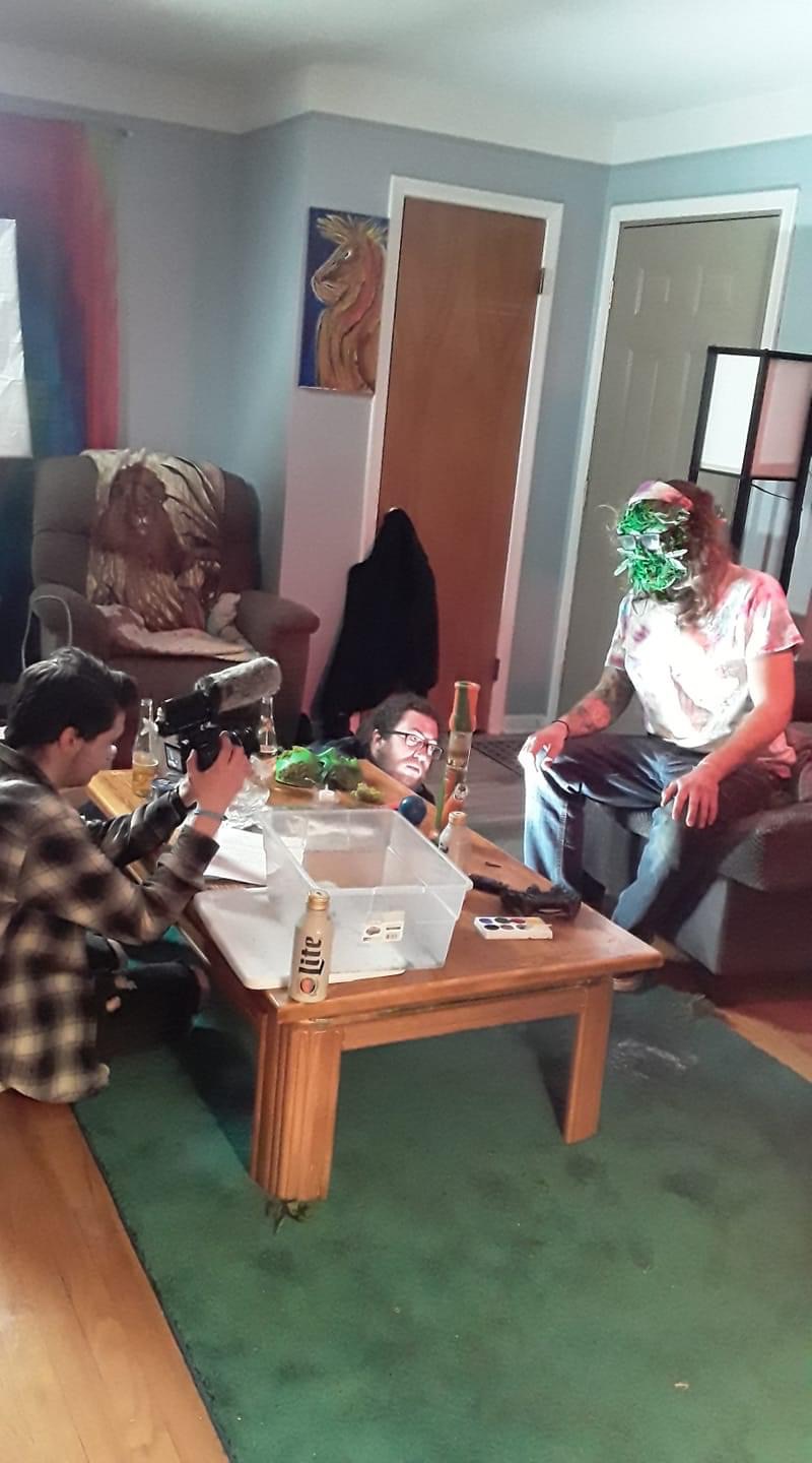 Person with green face paint sitting on a couch, another person taking photos, living room with furniture and artwork