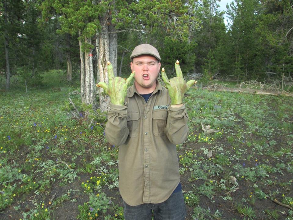 Person outdoors holding two large green gloves, trees and grass in background.