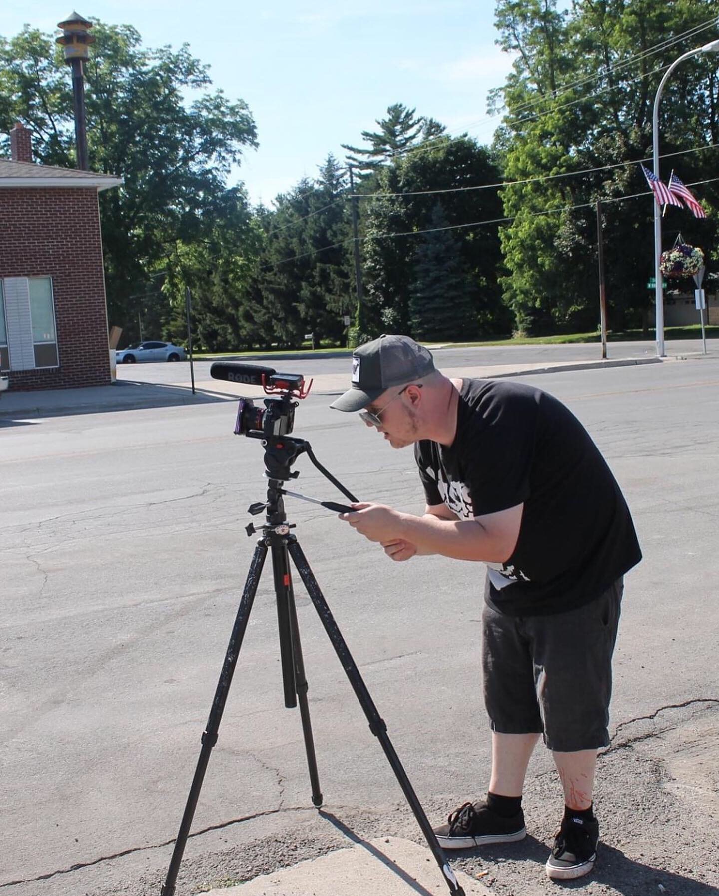 Young man with a cap and black t-shirt adjusting a camera on a tripod outdoors, with trees and a building in the background.