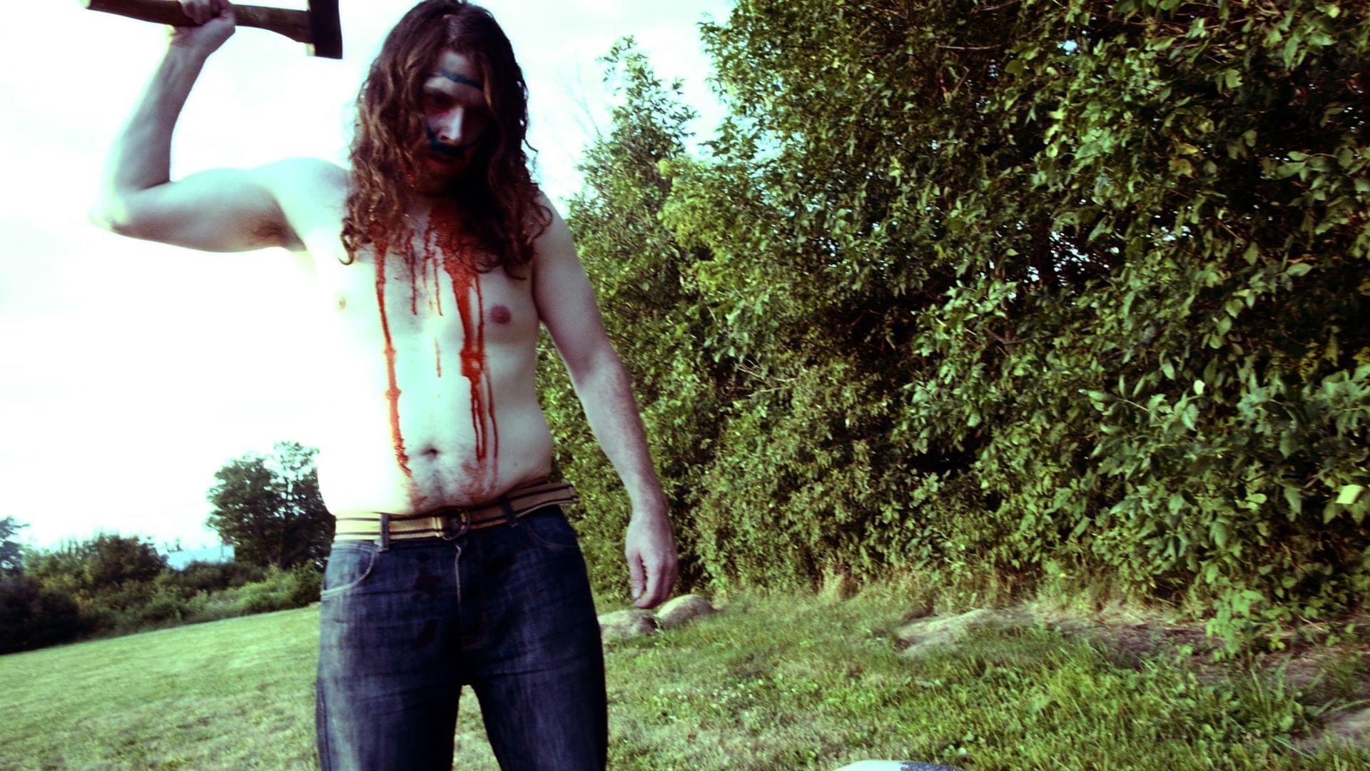 Person with long hair holding a weapon, standing outdoors near trees and grass.