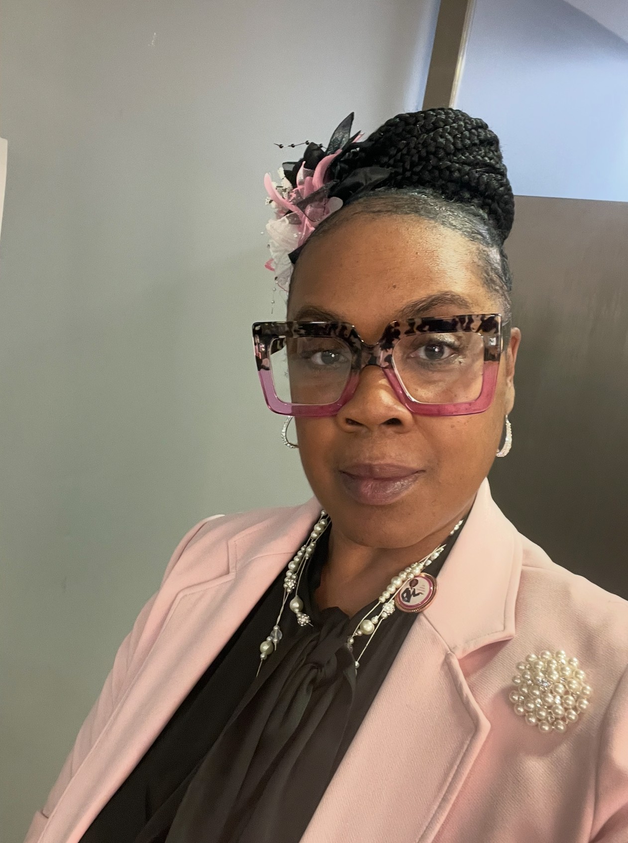 Woman with braided hair in a bun, wearing glasses, pink blazer, and pearl brooch, standing indoors.