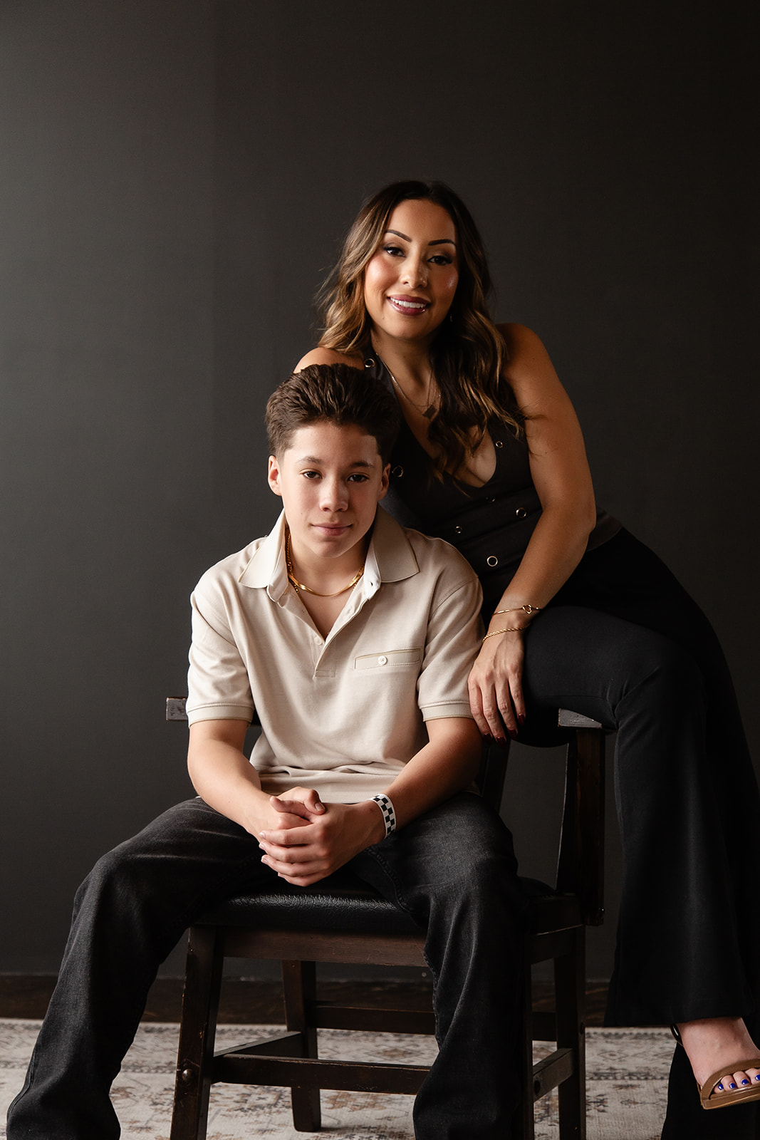A woman and a young boy sit together against a dark background, smiling at the camera. The woman has long wavy hair and wears a black dress. The boy wears a light-colored shirt and dark pants.