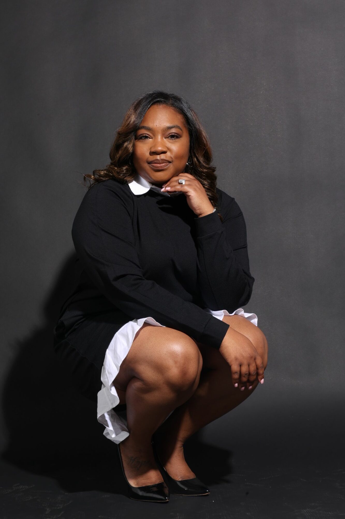 Woman sitting on the floor with legs crossed, smiling, wearing a black blazer and white shirt, dark background.