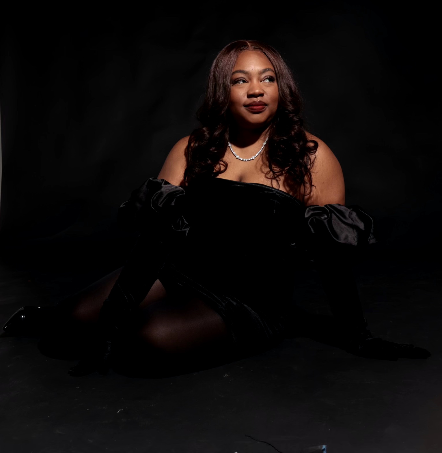 Woman with long hair and pearl necklace sitting against a dark background, wearing a black off-shoulder dress.