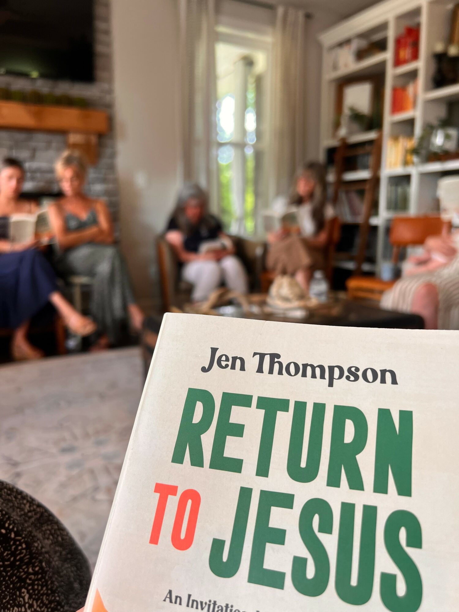 Book titled 'Return to Jesus' by Jen Thompson held in a living room with people sitting and talking, bookshelf, and window.
