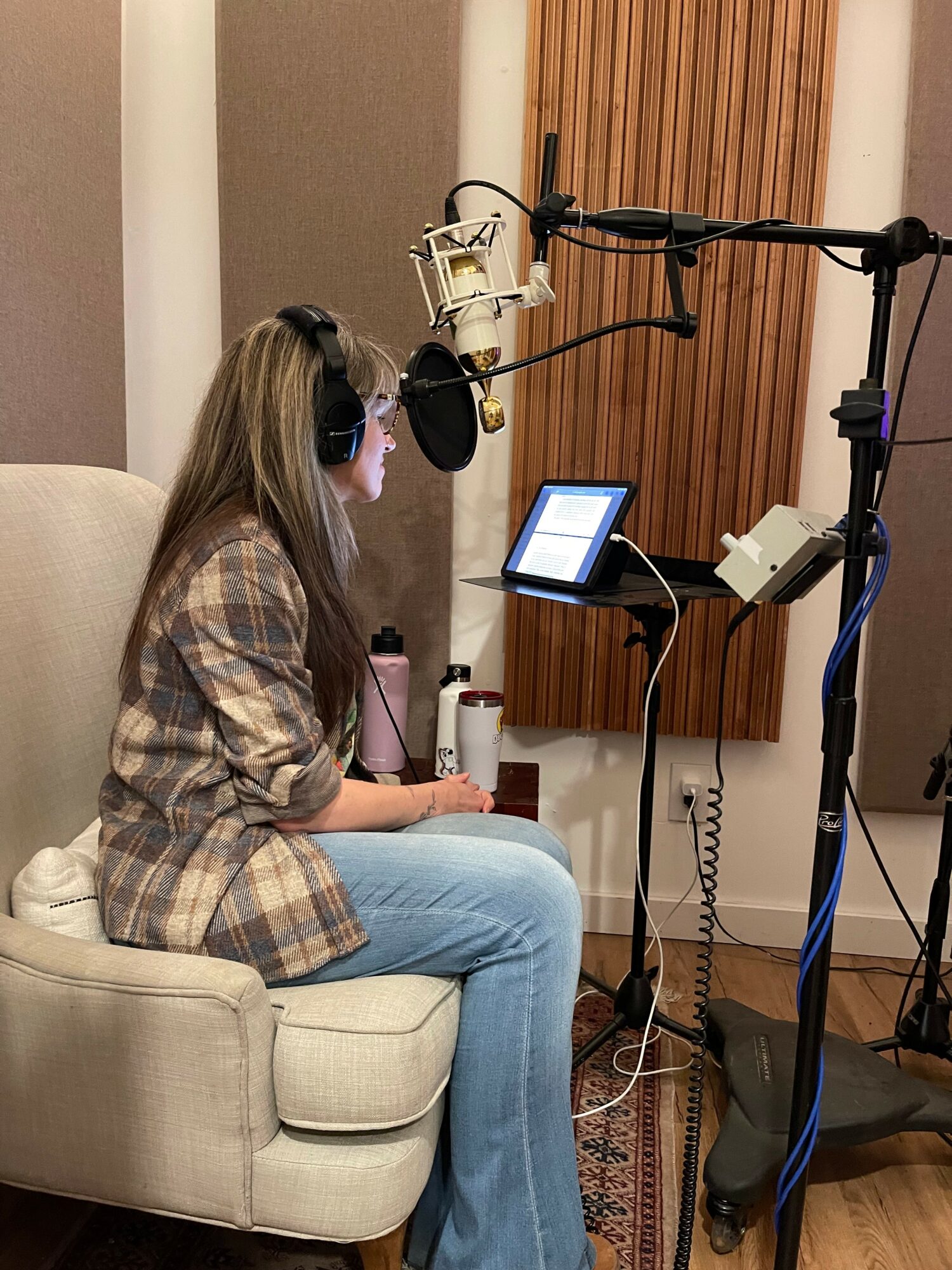 Woman sitting on a sofa in a recording studio, wearing headphones, with recording equipment and microphone setup nearby.
