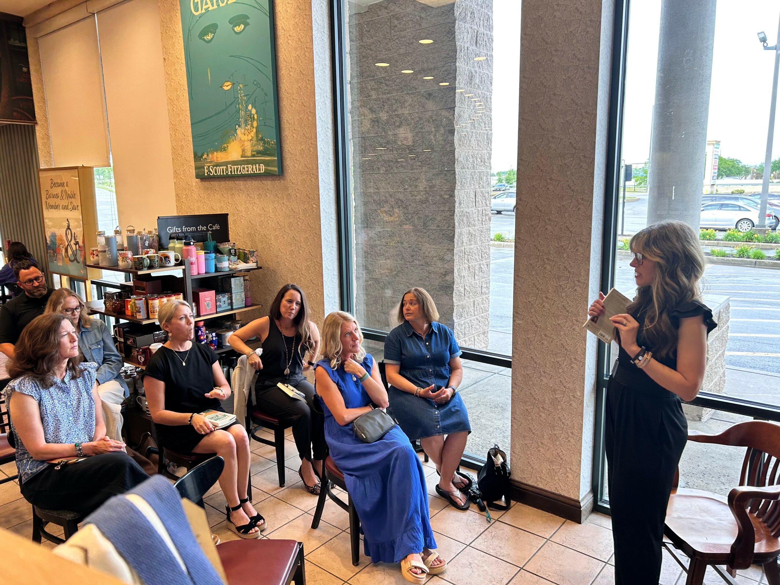 A woman stands and speaks to a seated group of women in a room with large windows and a view outside.