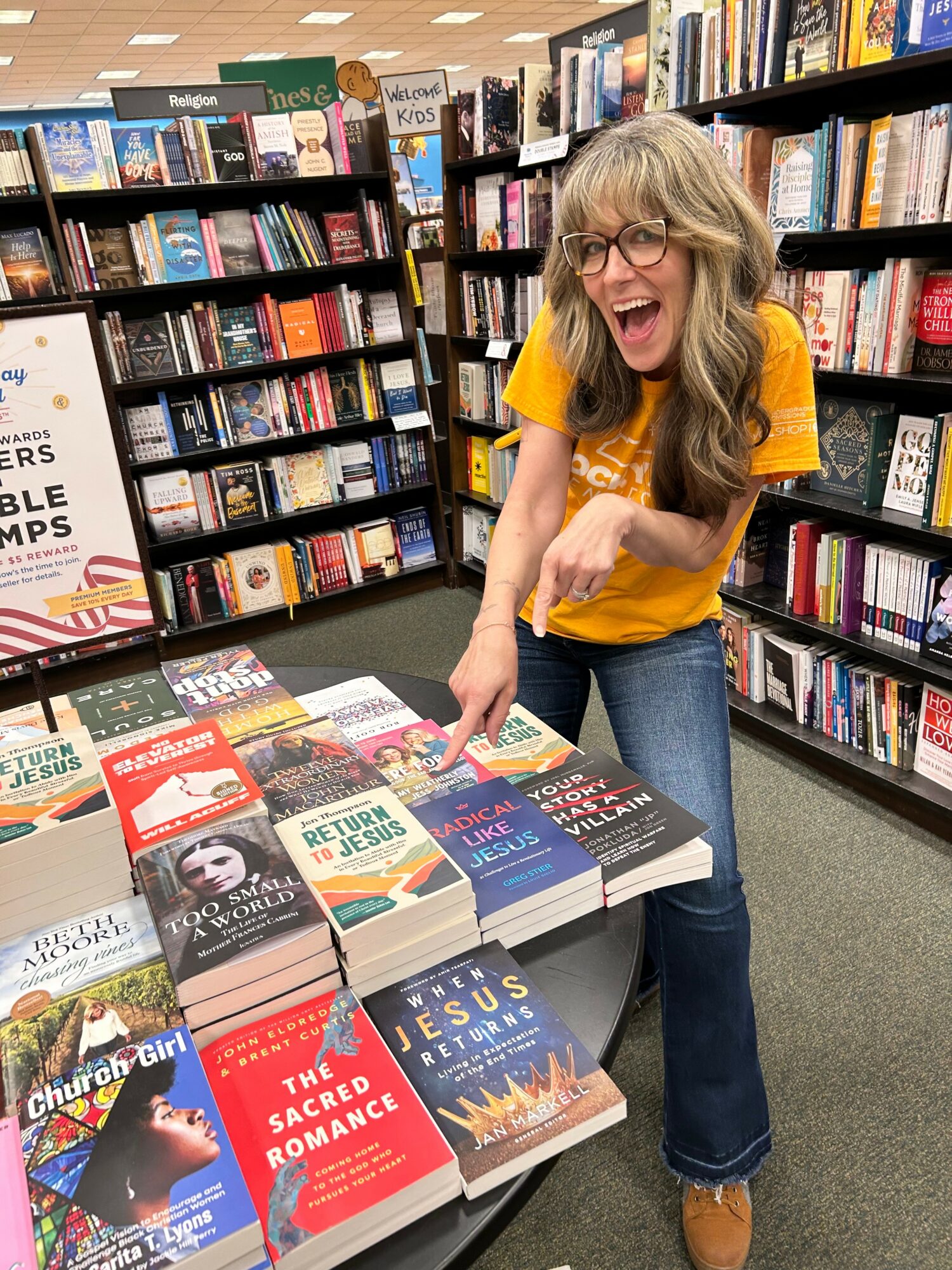 Woman with glasses and long hair, smiling and pointing at books on a table in a bookstore.