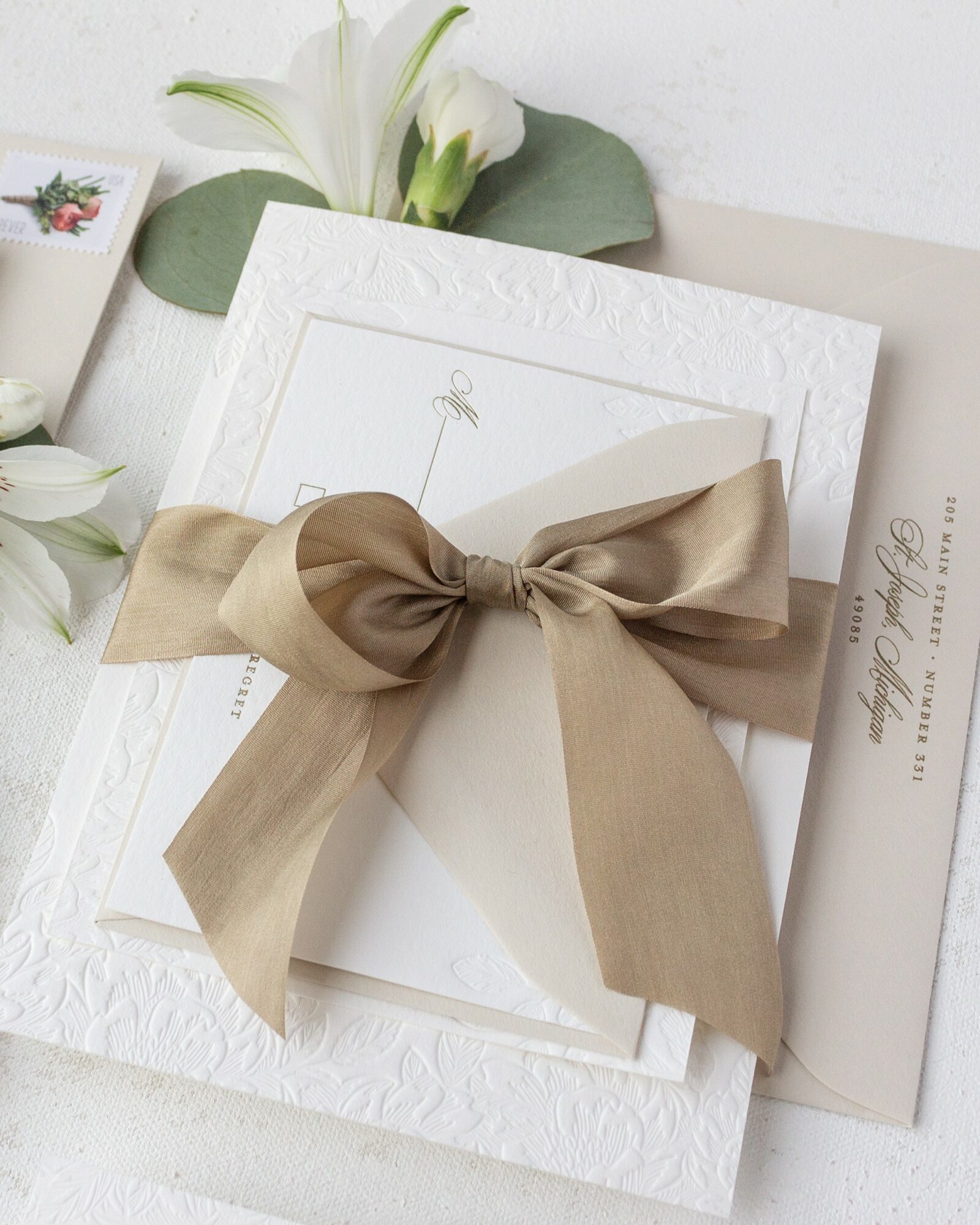 White textured card with a beige ribbon bow, surrounded by flowers and a paper with text, on a light surface.
