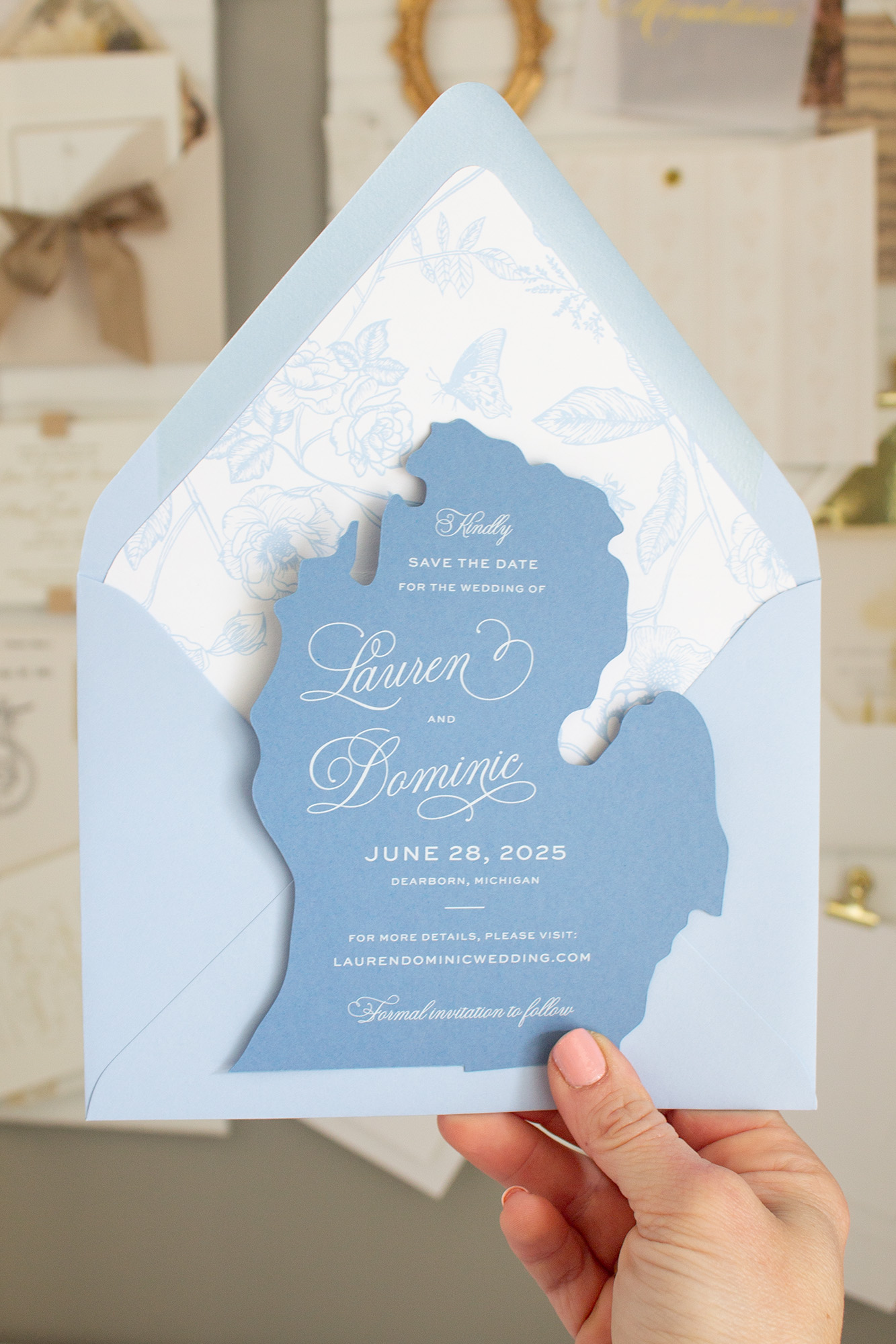 Hand holding a blue wedding invitation with a silhouette of a state, floral design, and event details.