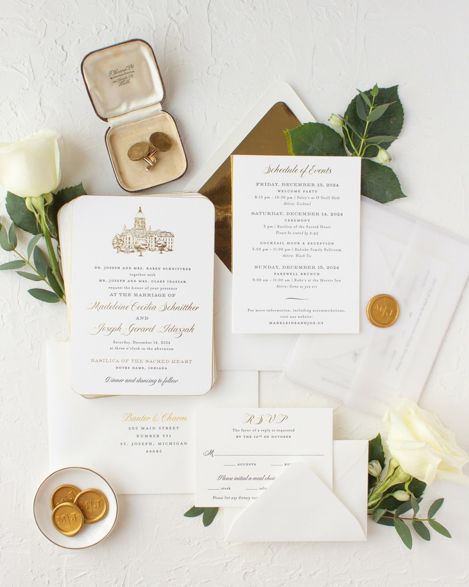 Wedding invitation, RSVP card, gold wax seal, flowers, and a ring box with rings on a white surface.