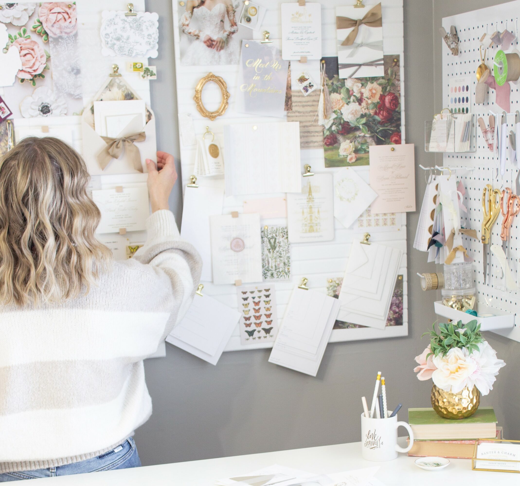 Person with wavy hair arranging items on a decorated bulletin board, with stationery and a flower pot on the desk.