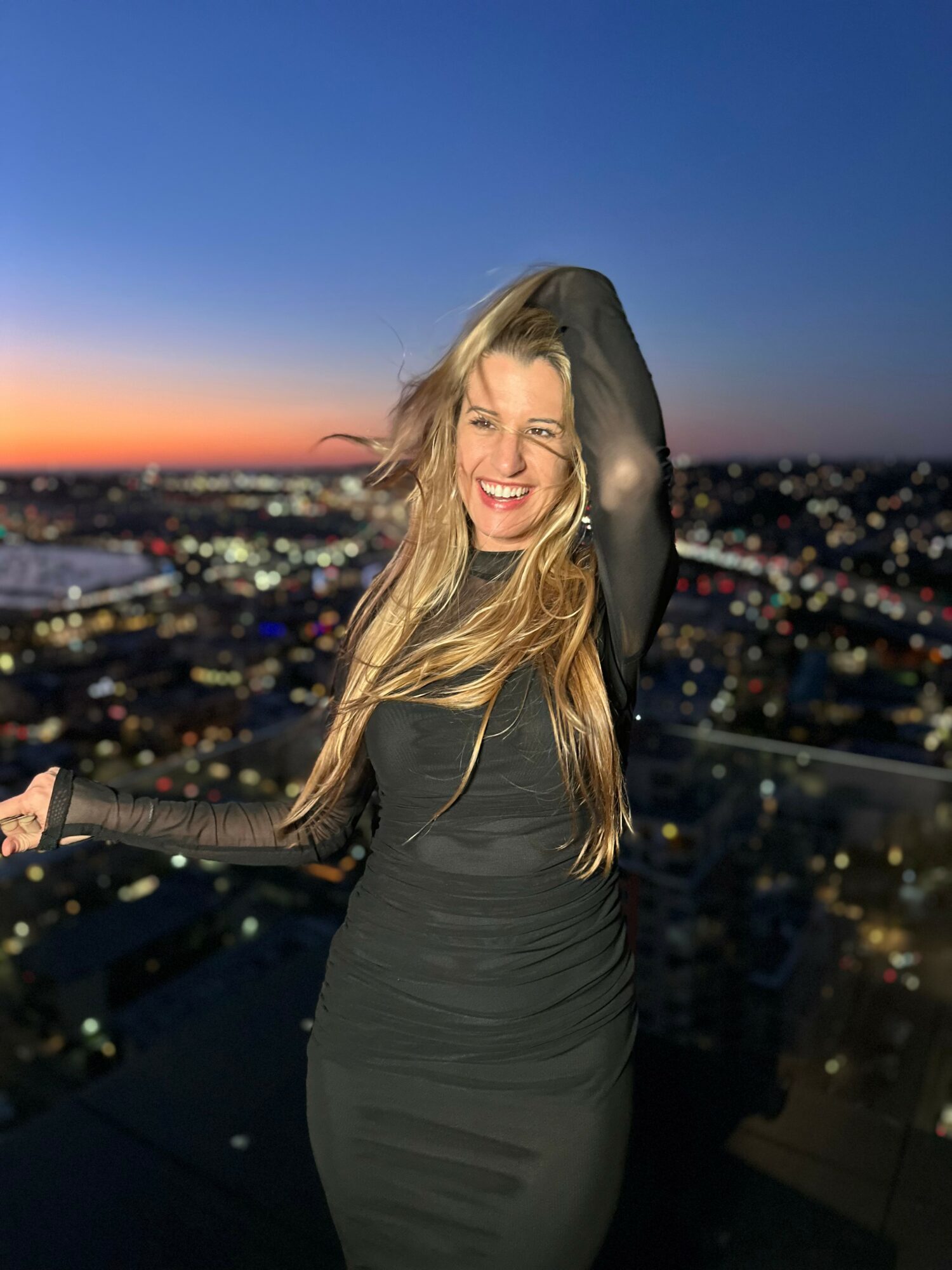 Woman with long hair smiling outdoors at sunset, cityscape in background, wearing a dark dress, hand on head.
