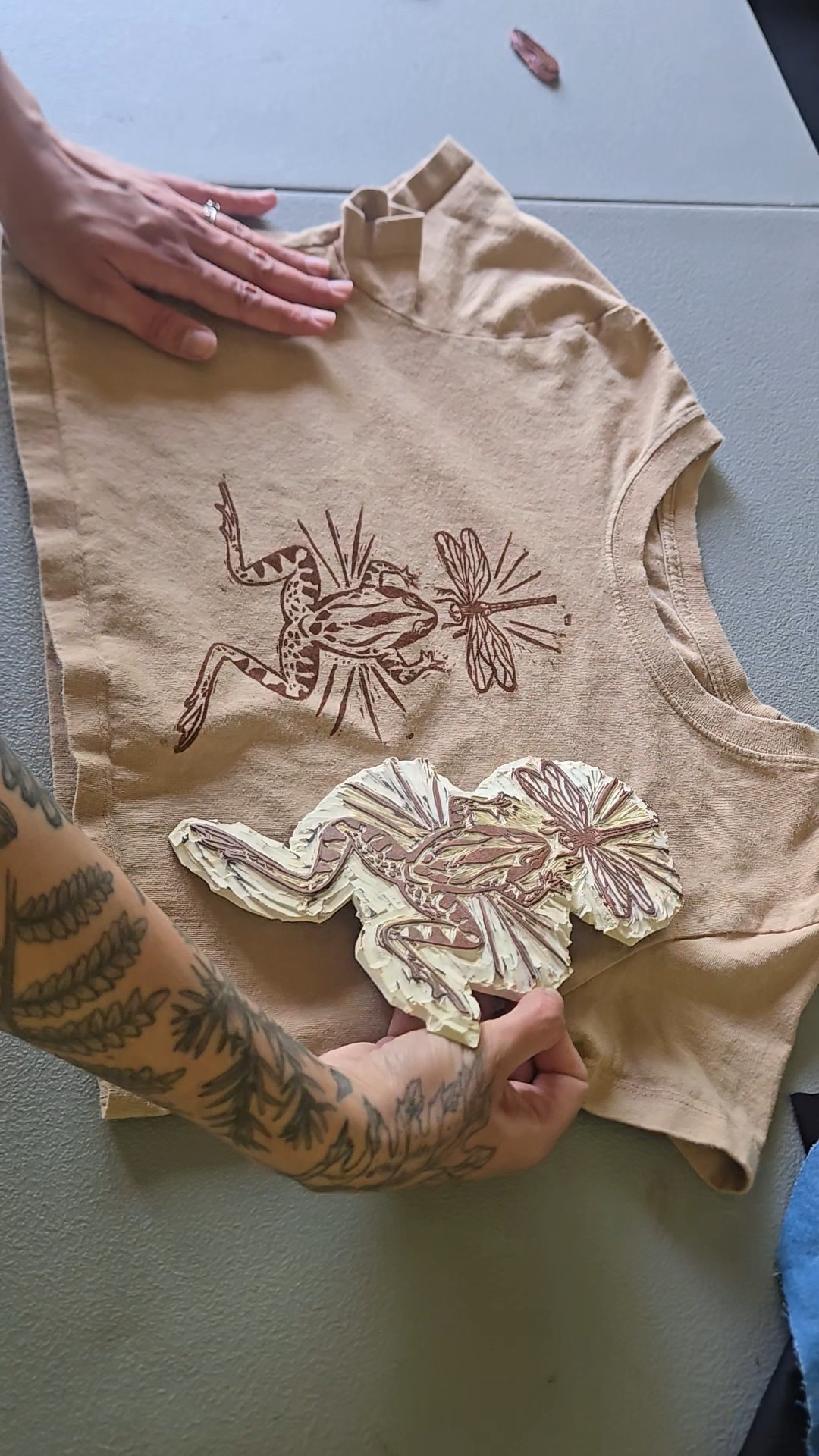 Person's hand and arm with tattoos holding a fabric piece with embroidered animal designs, on a table surface.