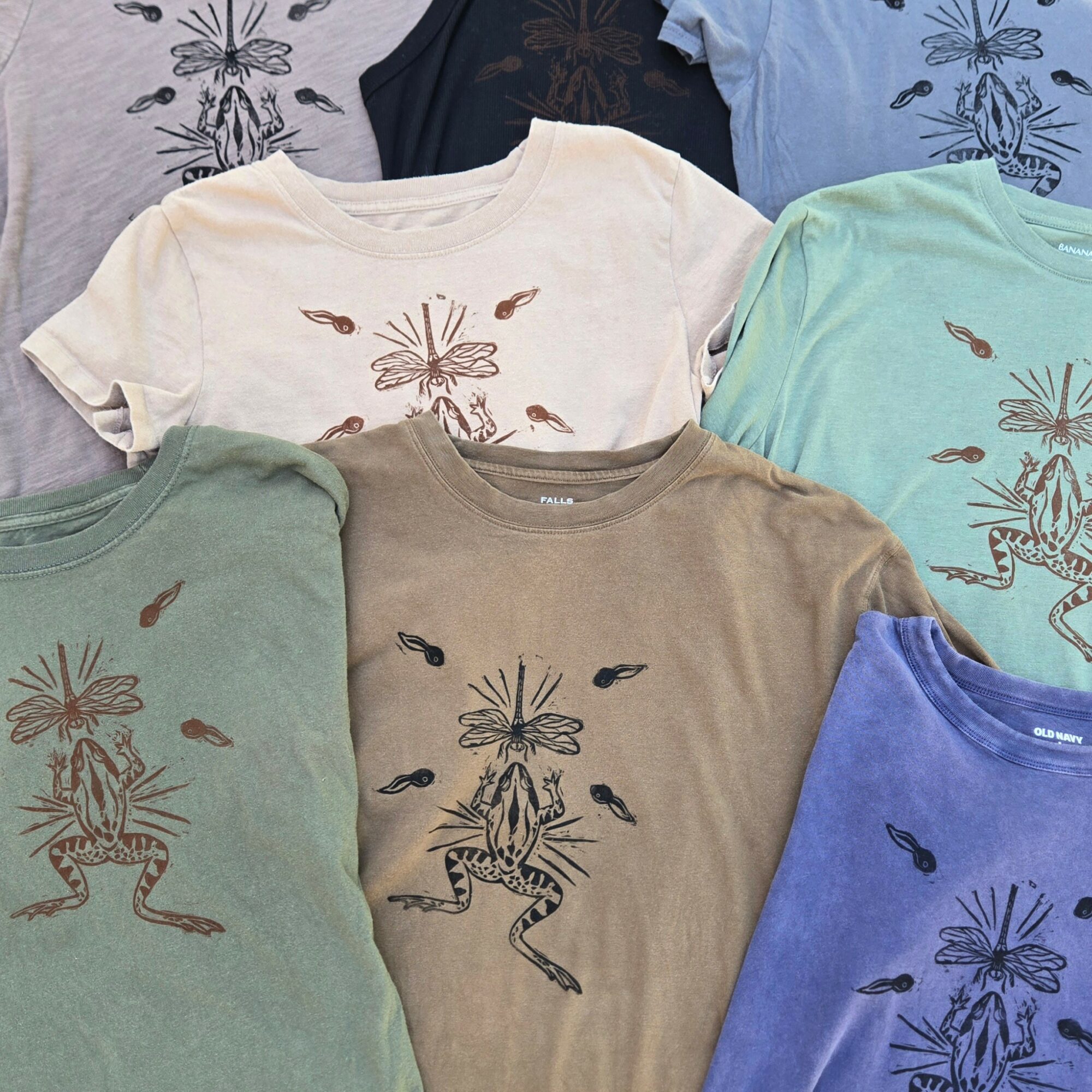 Six T-shirts with butterfly and frog designs, in various colors, overlapping each other.