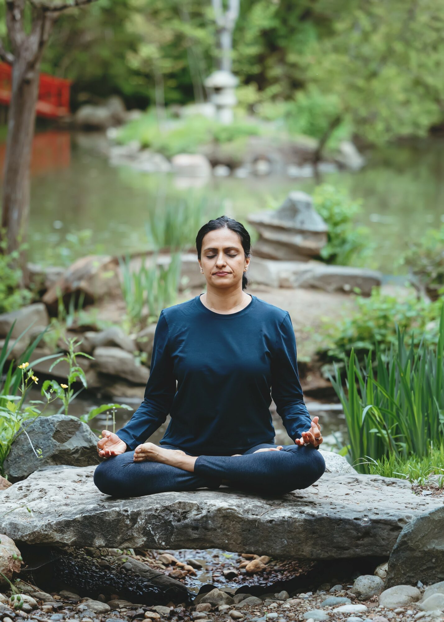 Person practicing meditation in a natural outdoor setting with greenery and water, sitting cross-legged on a rock.