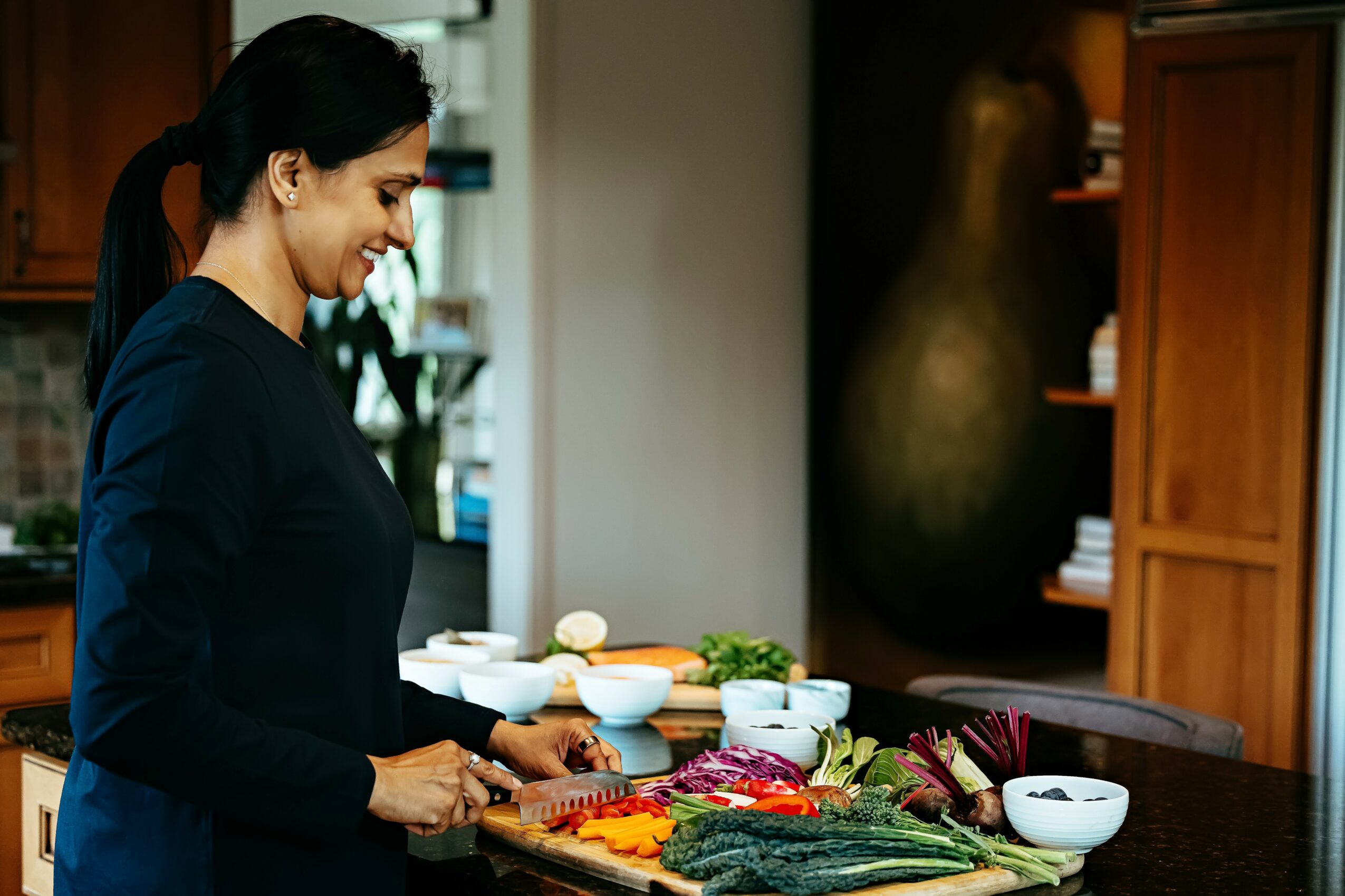 Woman smiling and preparing food in a kitchen with vegetables and bowls on the counter.