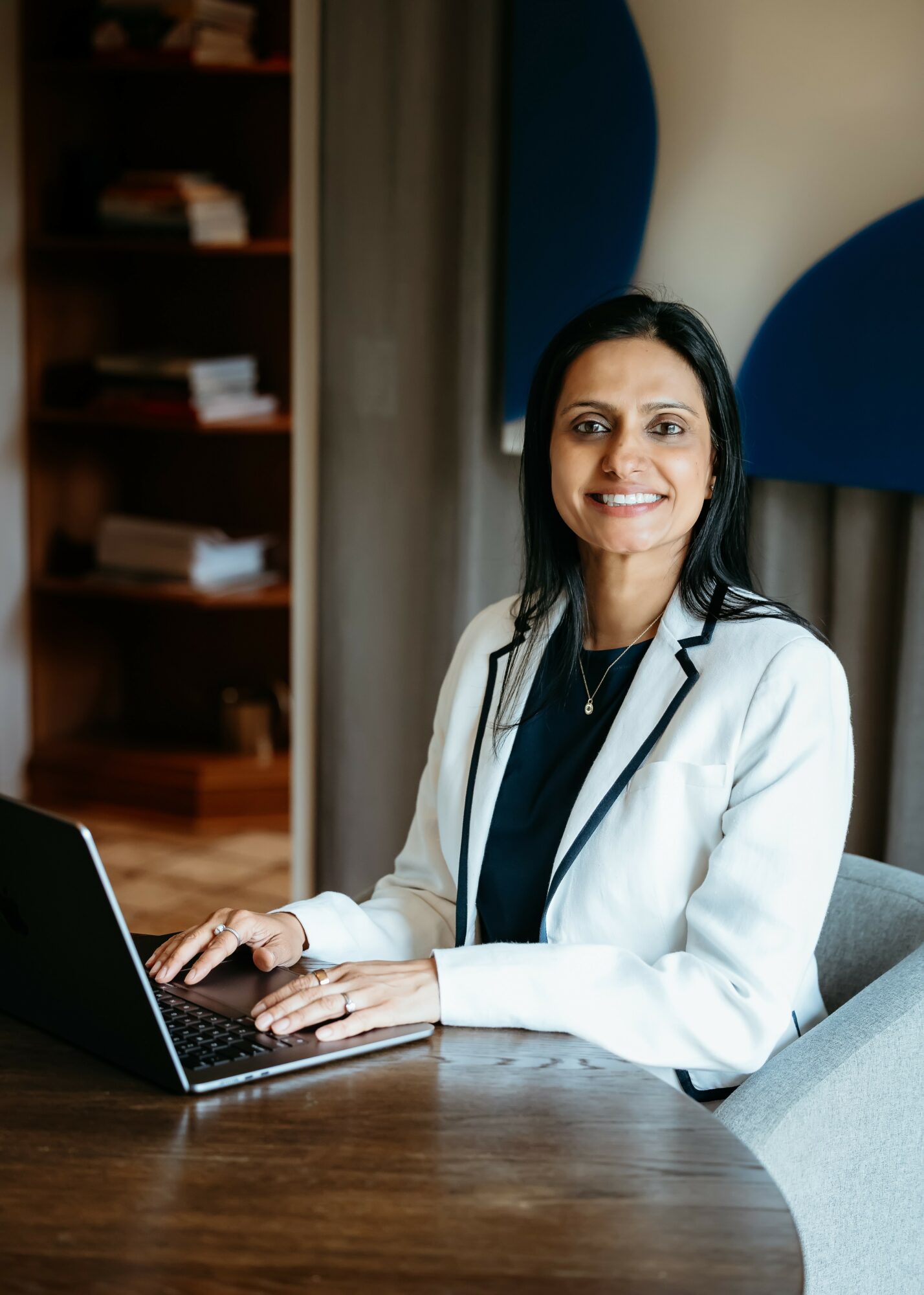 Woman in white blazer sitting at a desk with a laptop, smiling, in a room with bookshelves and curtains.