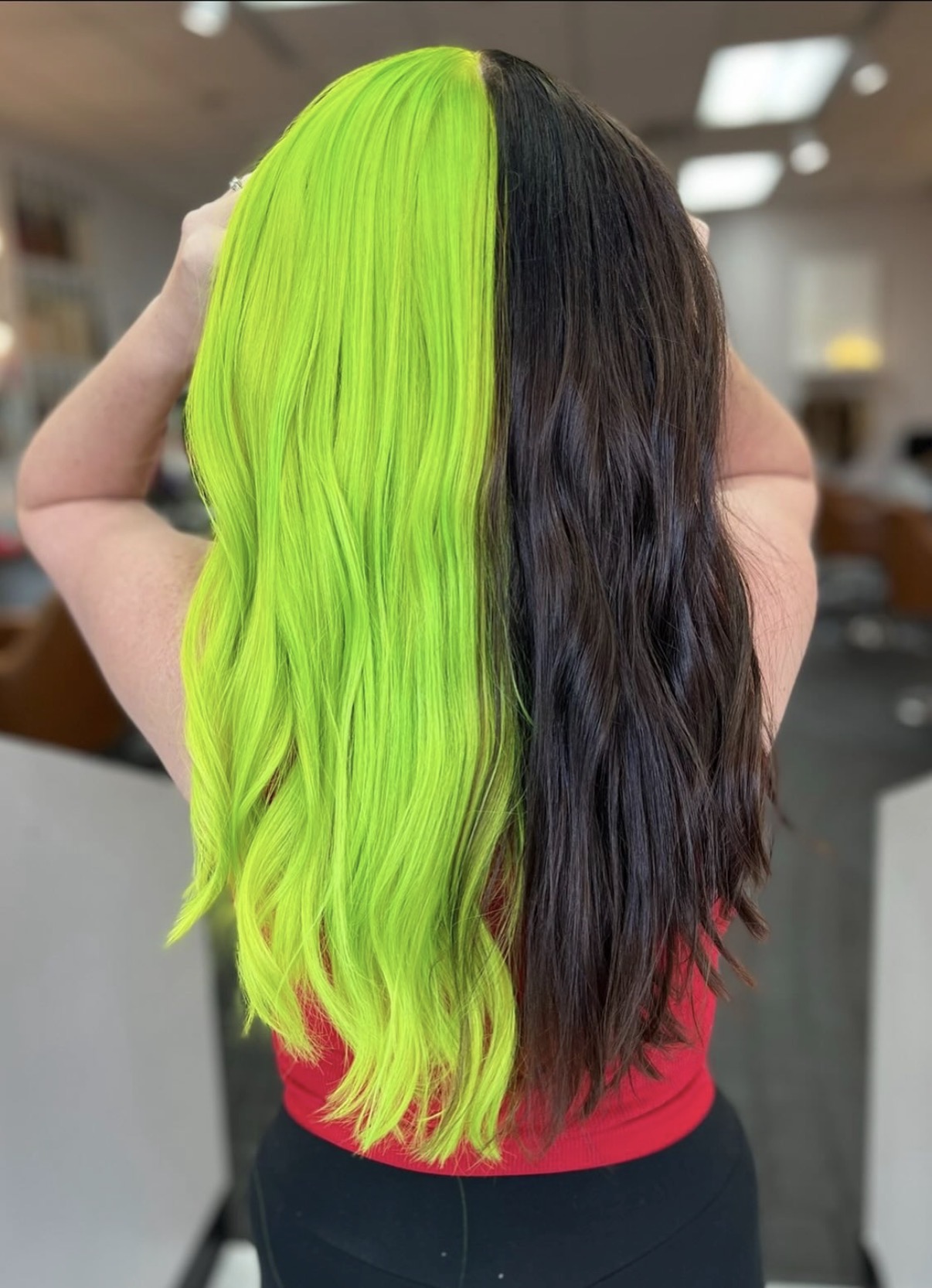 Back view of a person with half bright green and half dark brown wavy hair, wearing a red top.
