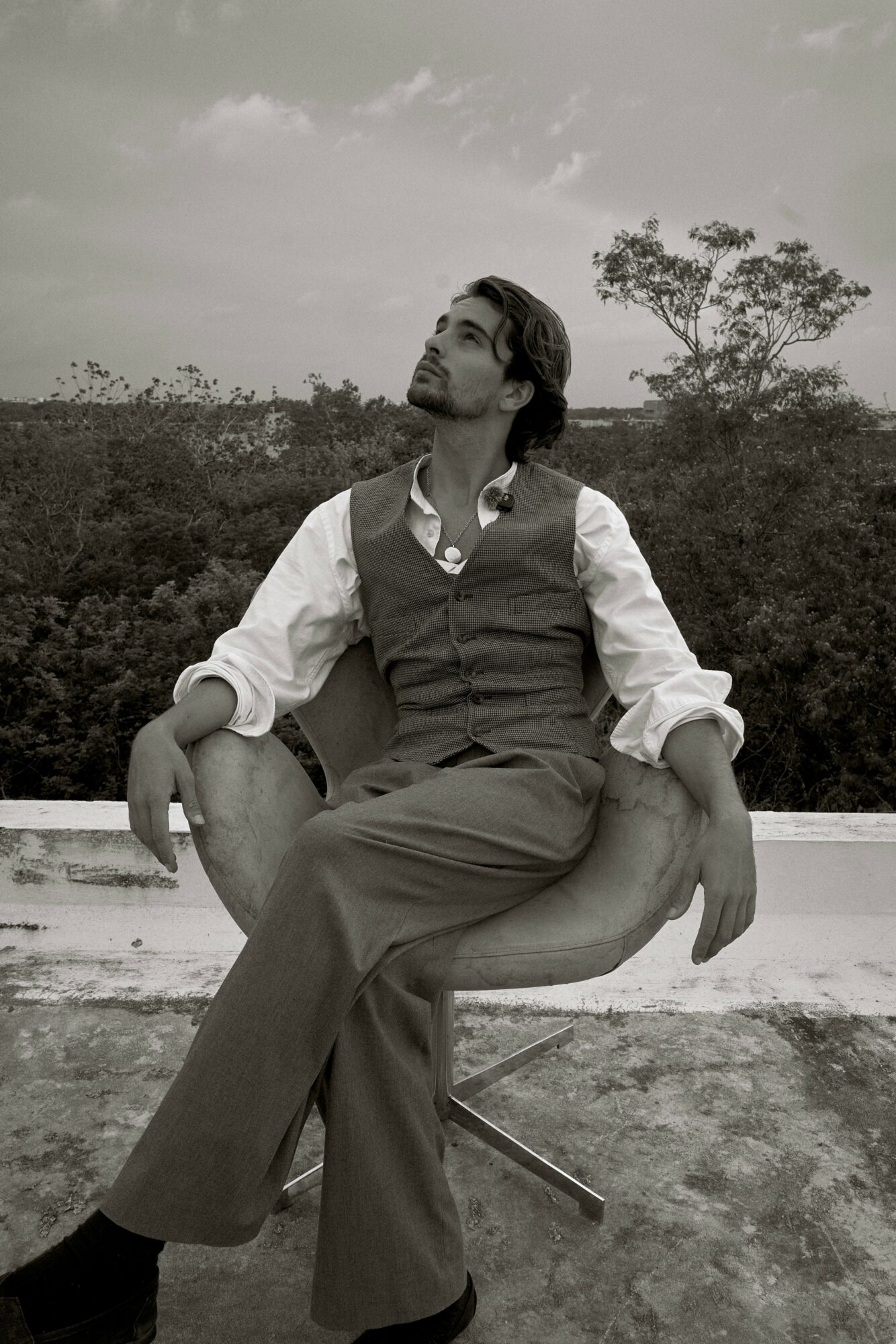 Person sitting on a chair outdoors, looking up, with trees and sky in background, in black and white.