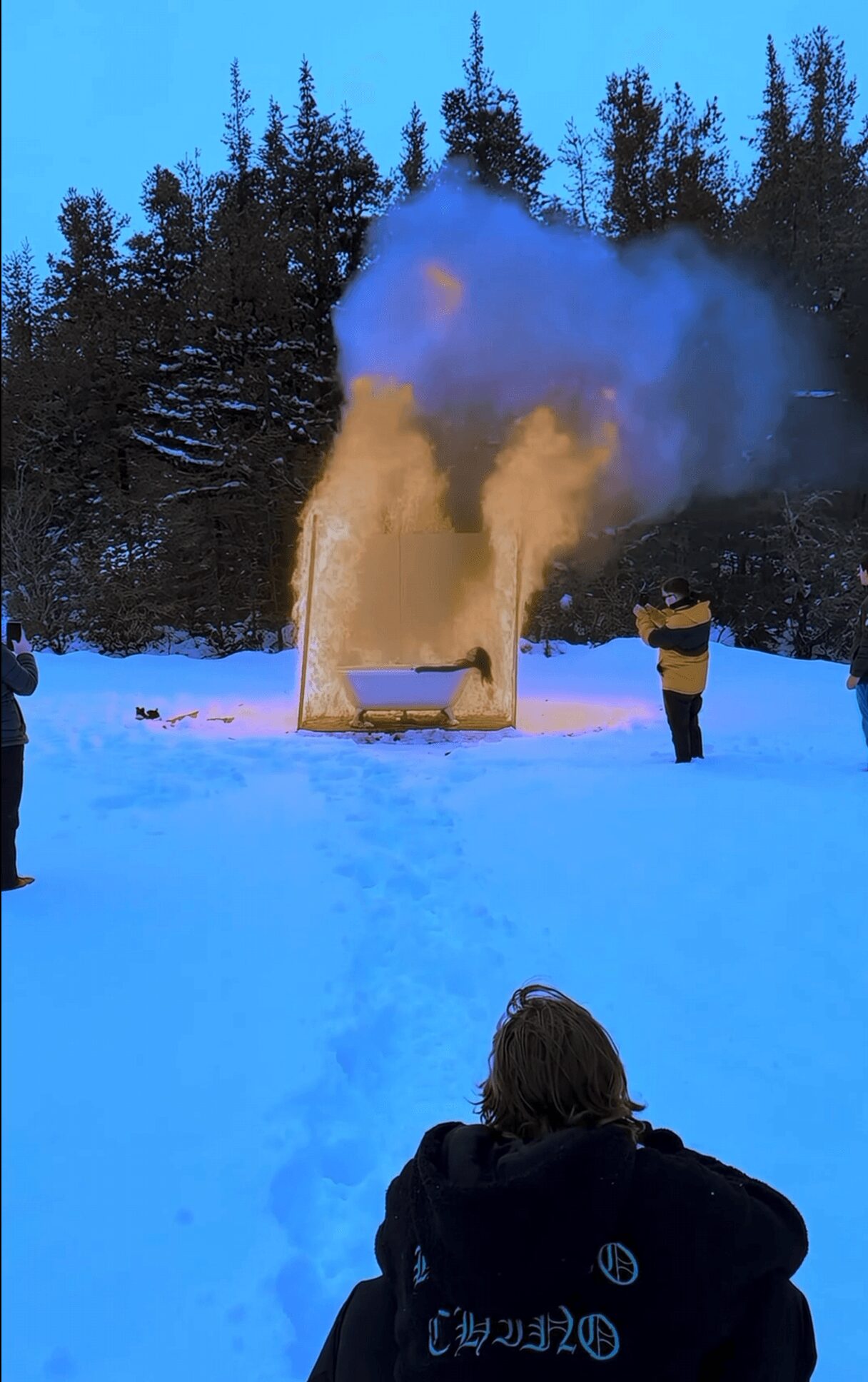 Person in dark clothing watches a steam or smoke cloud rising from a yellow object in snowy landscape with trees in background.