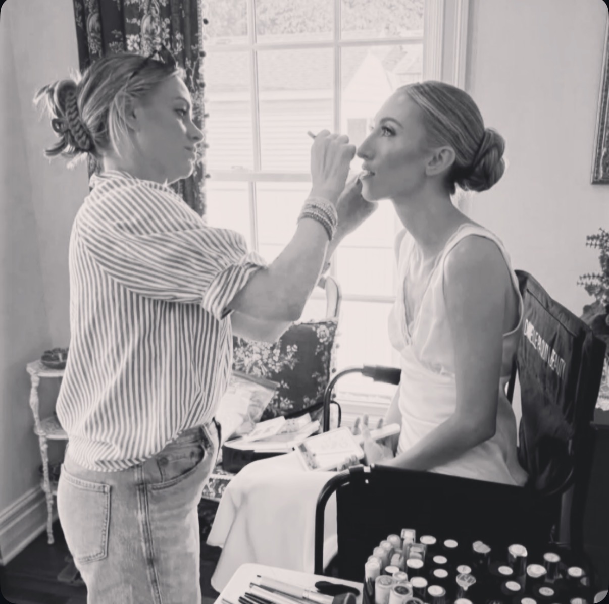 Makeup artist applies lipstick to a woman sitting in a chair indoors, with a window in the background.