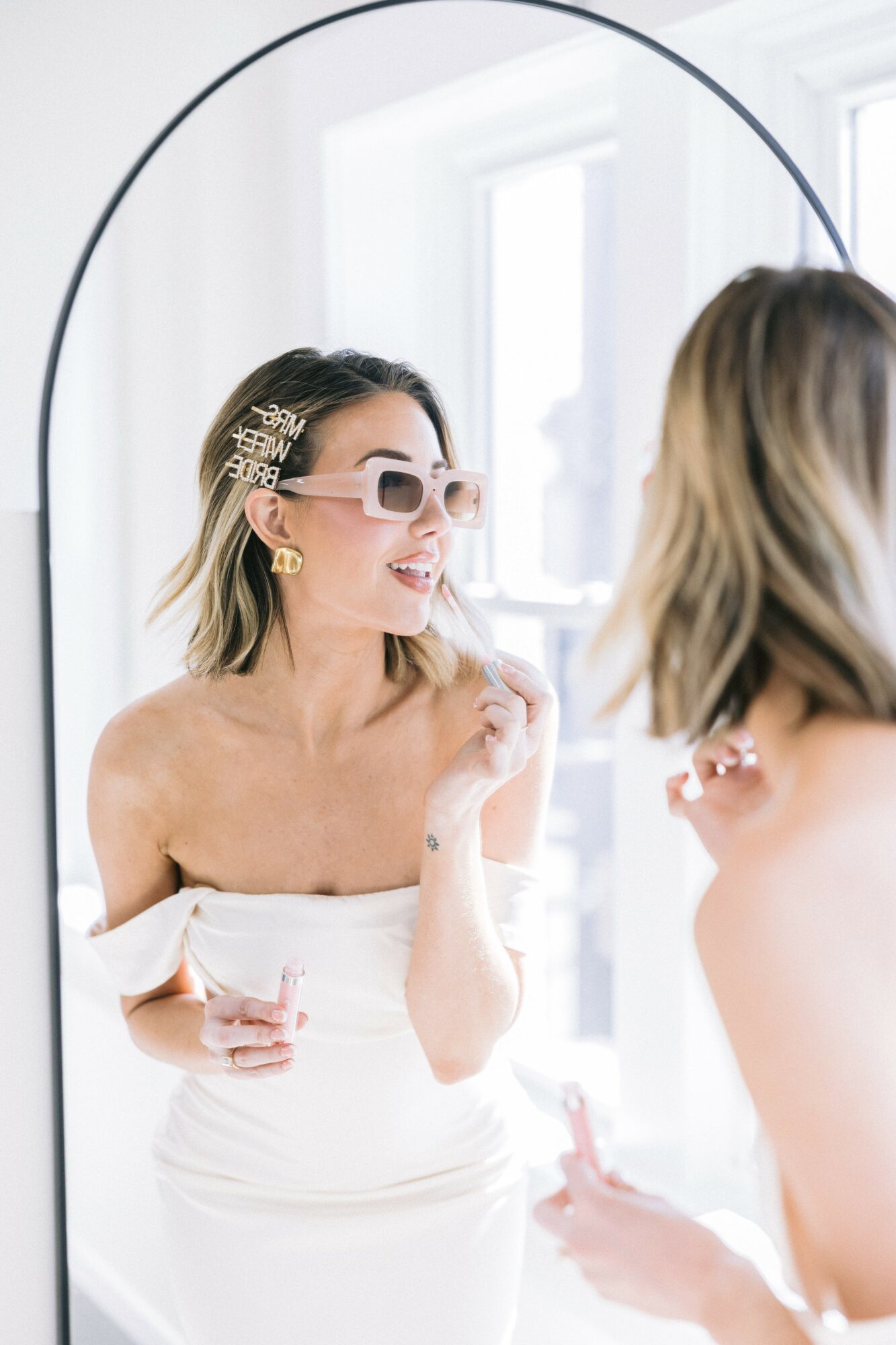 Woman with sunglasses and hair clips looking at her reflection in a mirror, smiling, in a bright room.