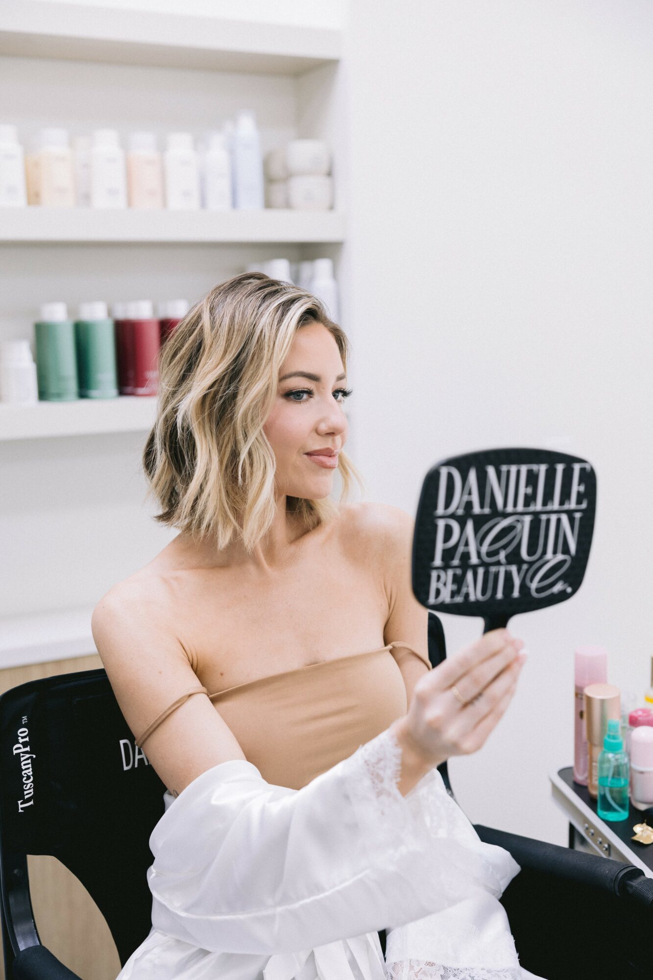 Woman with wavy blonde hair holding a sign that says 'DANIELLE PAGAN BEAUTY' in a salon, shelves with hair products in background.
