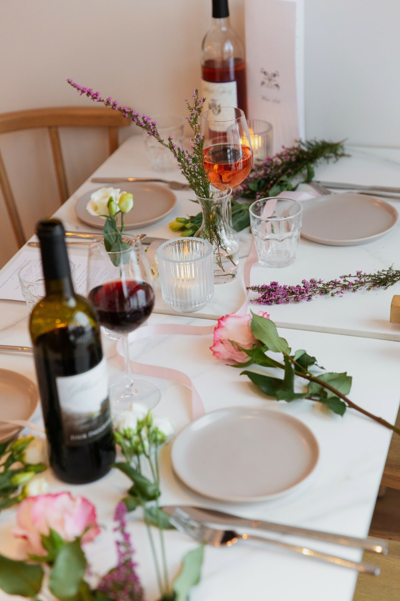 Table set with wine bottles, glasses, plates, flowers, and a bottle of rosé wine, decorated with greenery and flowers.