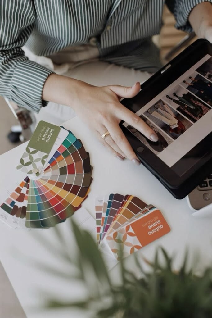 Person using a tablet with color swatches and fabric samples on a table, blurred plant in foreground.