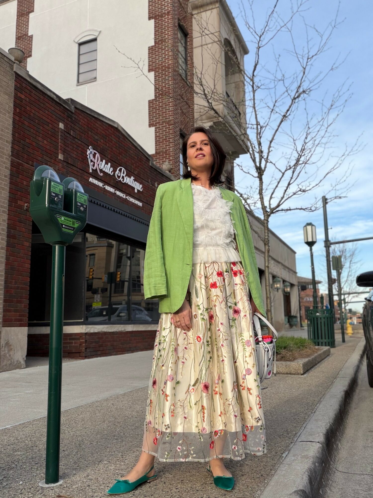 Woman in green jacket and floral skirt stands on city sidewalk with buildings and leafless trees behind her.