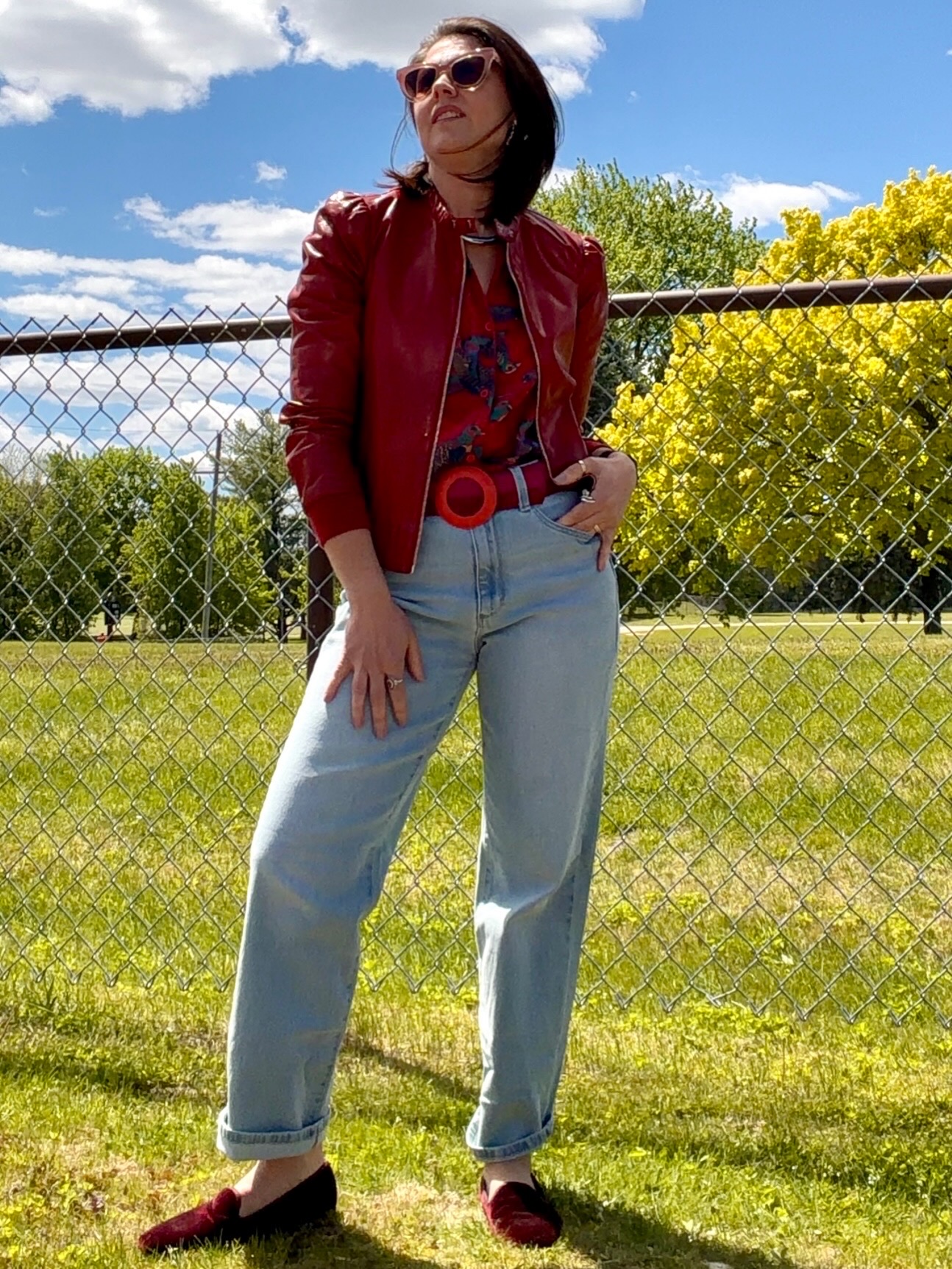 Woman standing outdoors in front of a chain-link fence, wearing sunglasses, a red jacket, light jeans, and black shoes.