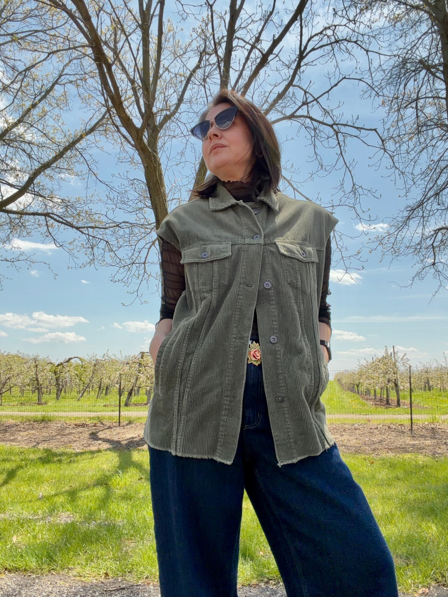 Woman standing outdoors in a grassy area with trees, wearing sunglasses, a green vest, and dark pants.