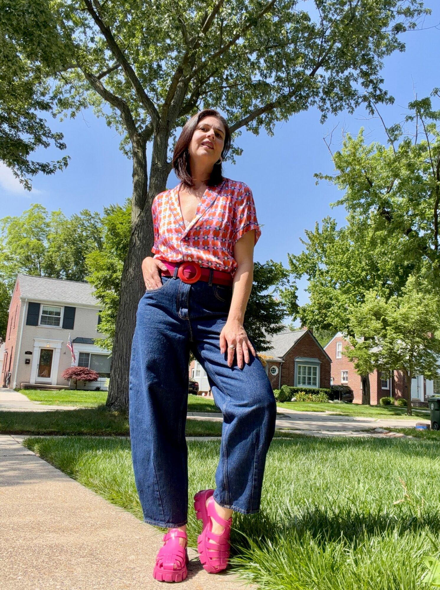 Young woman standing outdoors on a sidewalk, wearing a pink patterned top, high-waisted jeans, and pink shoes, with trees and houses in background.