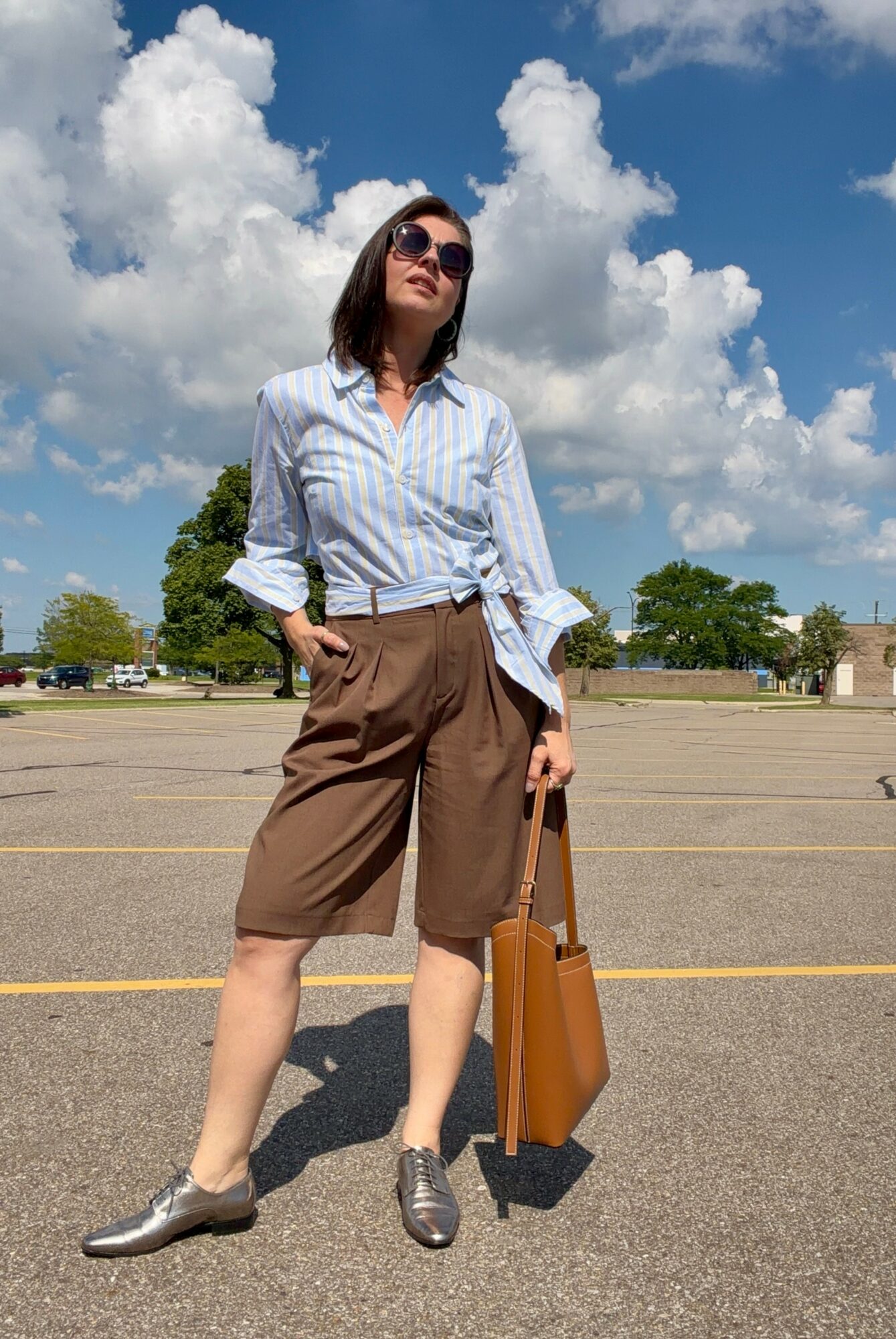 Woman standing in an empty parking lot with clouds and trees in the background, wearing sunglasses, a striped shirt, and brown shorts, holding a large bag.