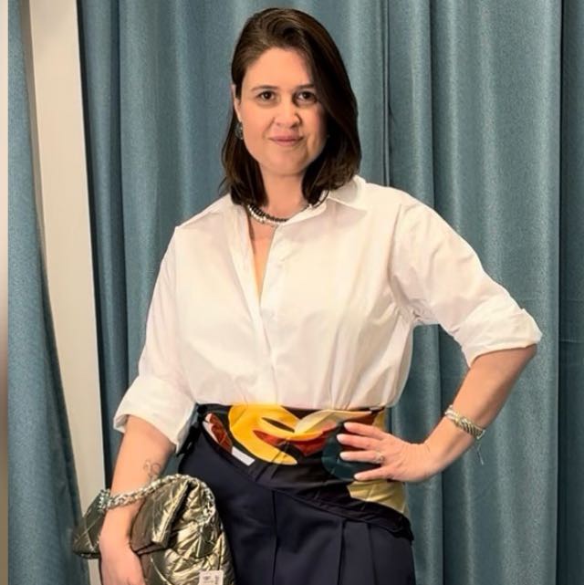 Woman with shoulder-length dark hair wearing a white shirt, black pants, and jewelry, standing in front of blue curtains.
