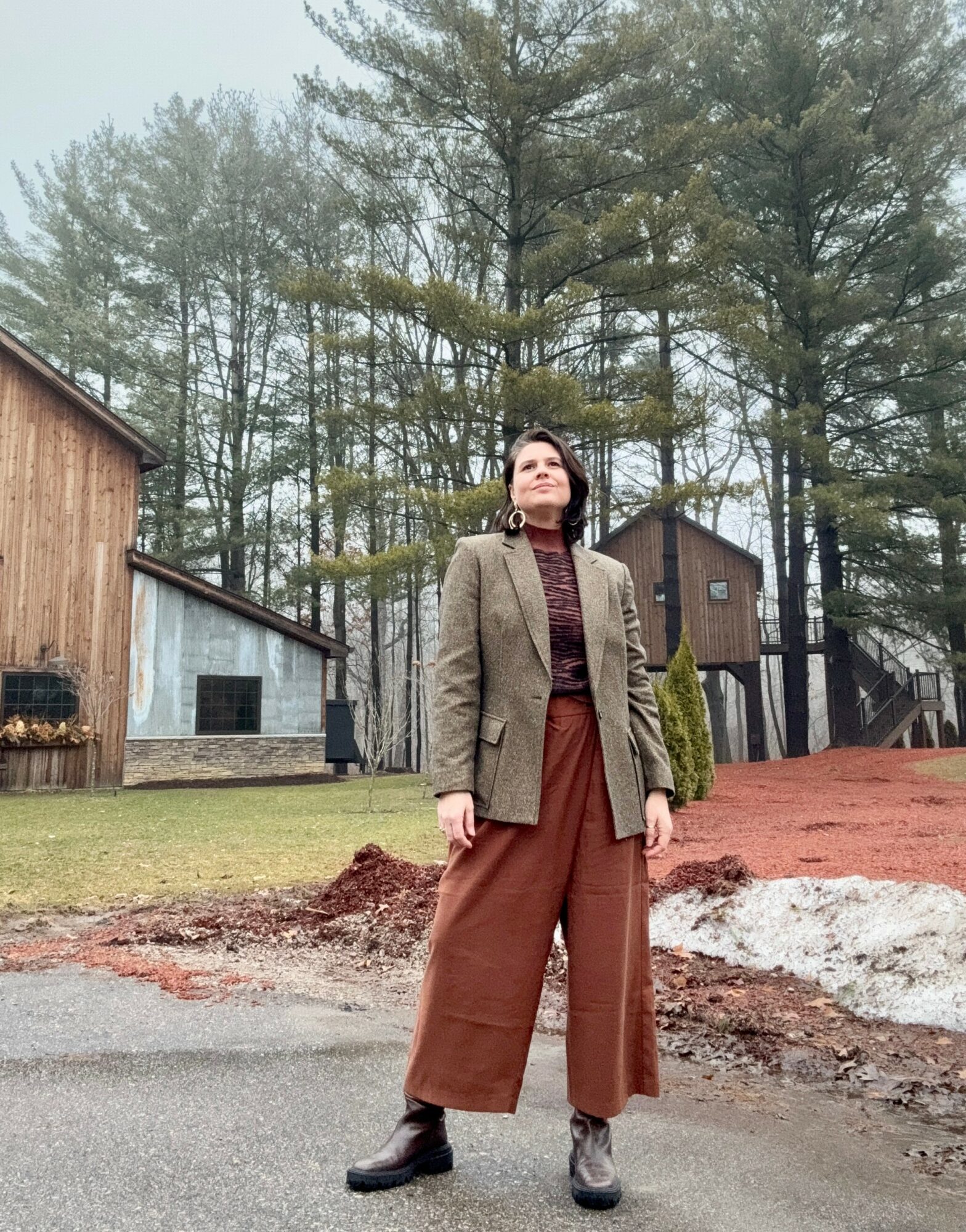Woman standing outdoors in a wooded area, wearing a blazer, turtleneck, wide pants, and boots, looking upward.